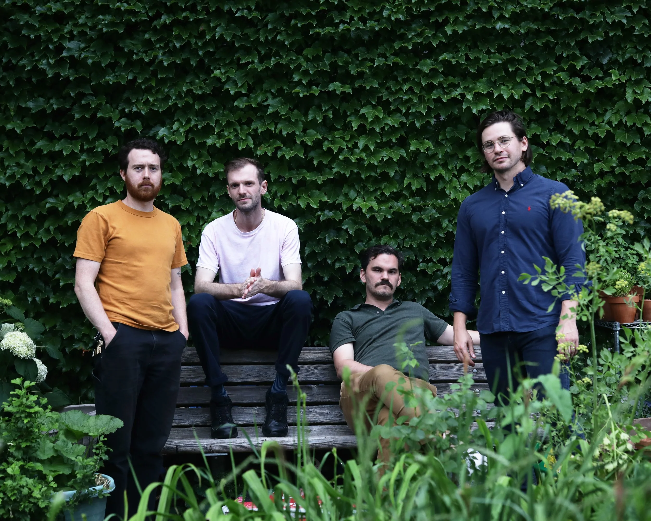 Five men outdoors in front of a wall of ivy, some sitting and some standing, surrounded by plants in pots.