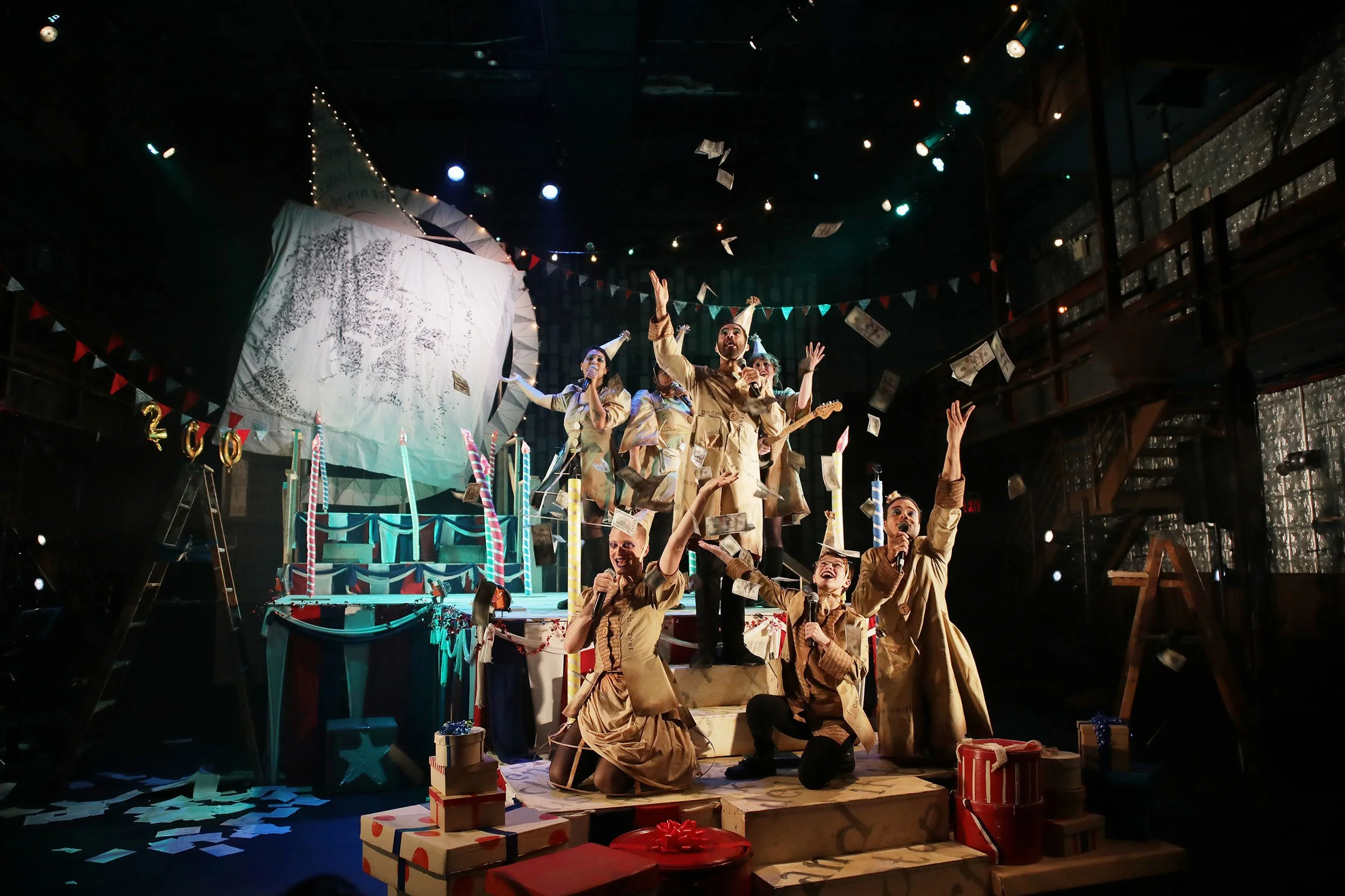 The image shows a lively stage scene with people in costumes celebrating. There are performers, some holding microphones, surrounded by Christmas decorations, presents, and confetti, indicating a festive holiday celebration.