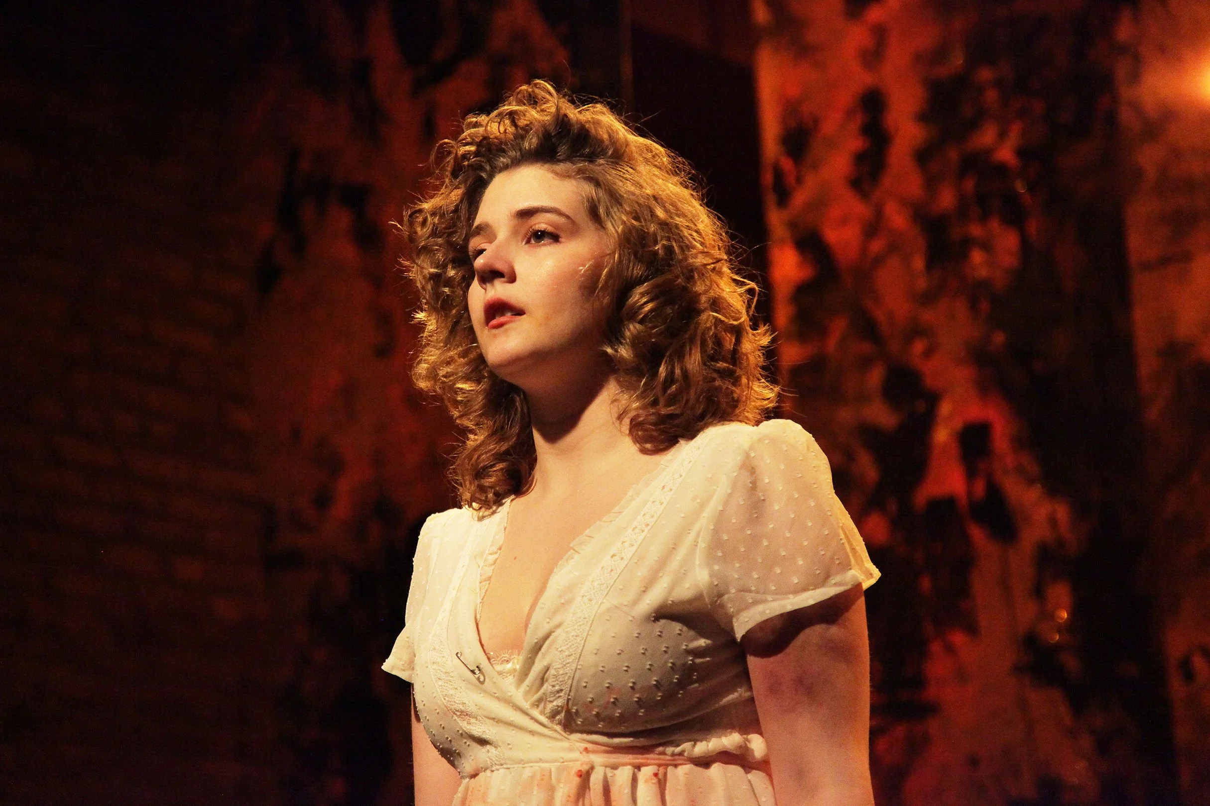 A woman with curly hair wearing a light-colored dress standing against a dark, textured background.