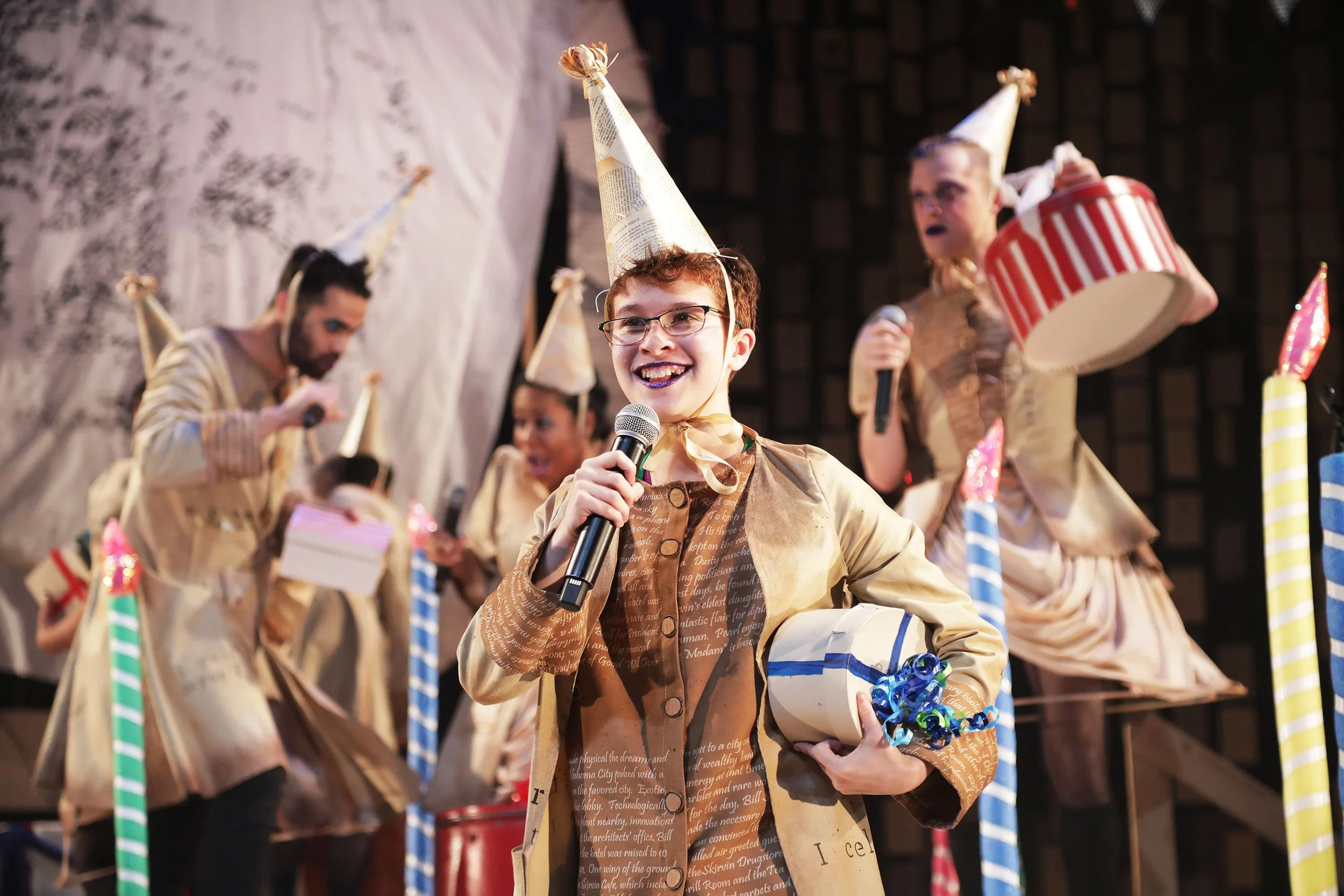 People wearing party hats and costumes at an indoor celebration, with a person in the foreground holding a microphone and balloons.