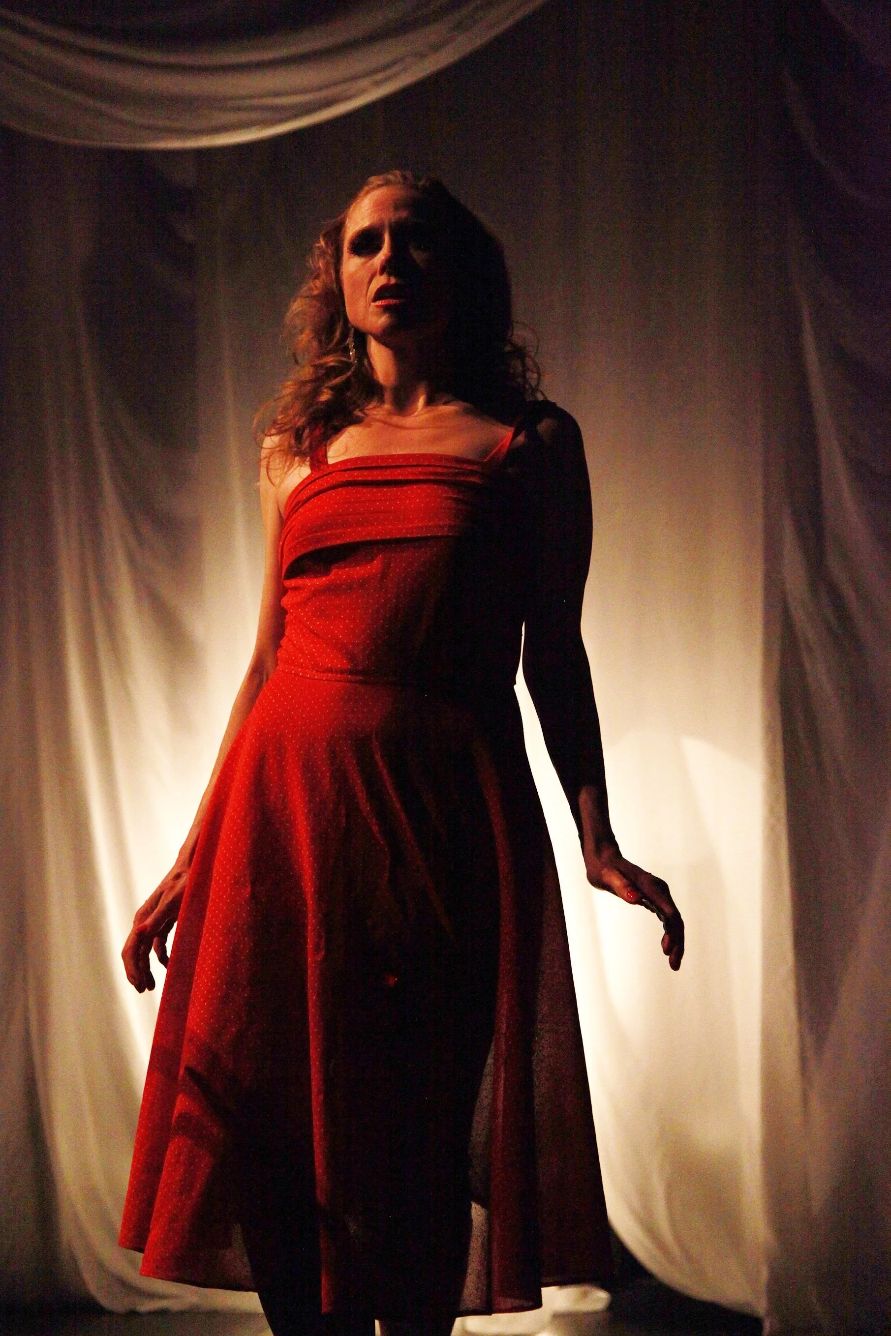 A woman with wavy hair wearing a red dress on a stage with dim lighting and a curtain backdrop.