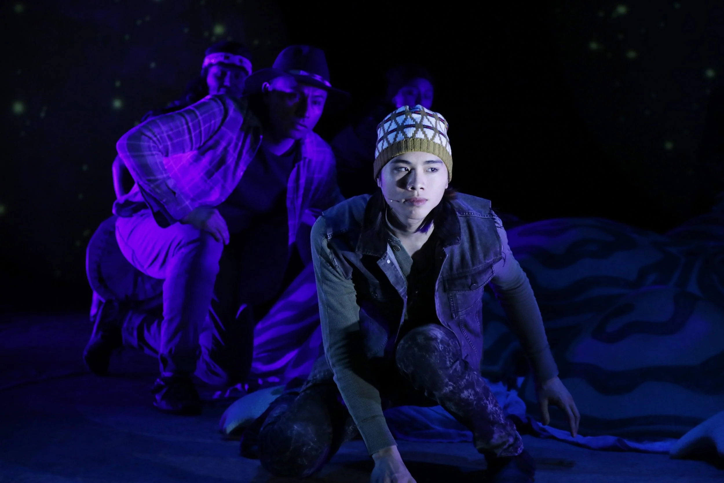 Four young people wearing colorful hats and casual clothing, sitting or crouching on a dark stage with black background and green spots of light, illuminated by purple and blue lights.