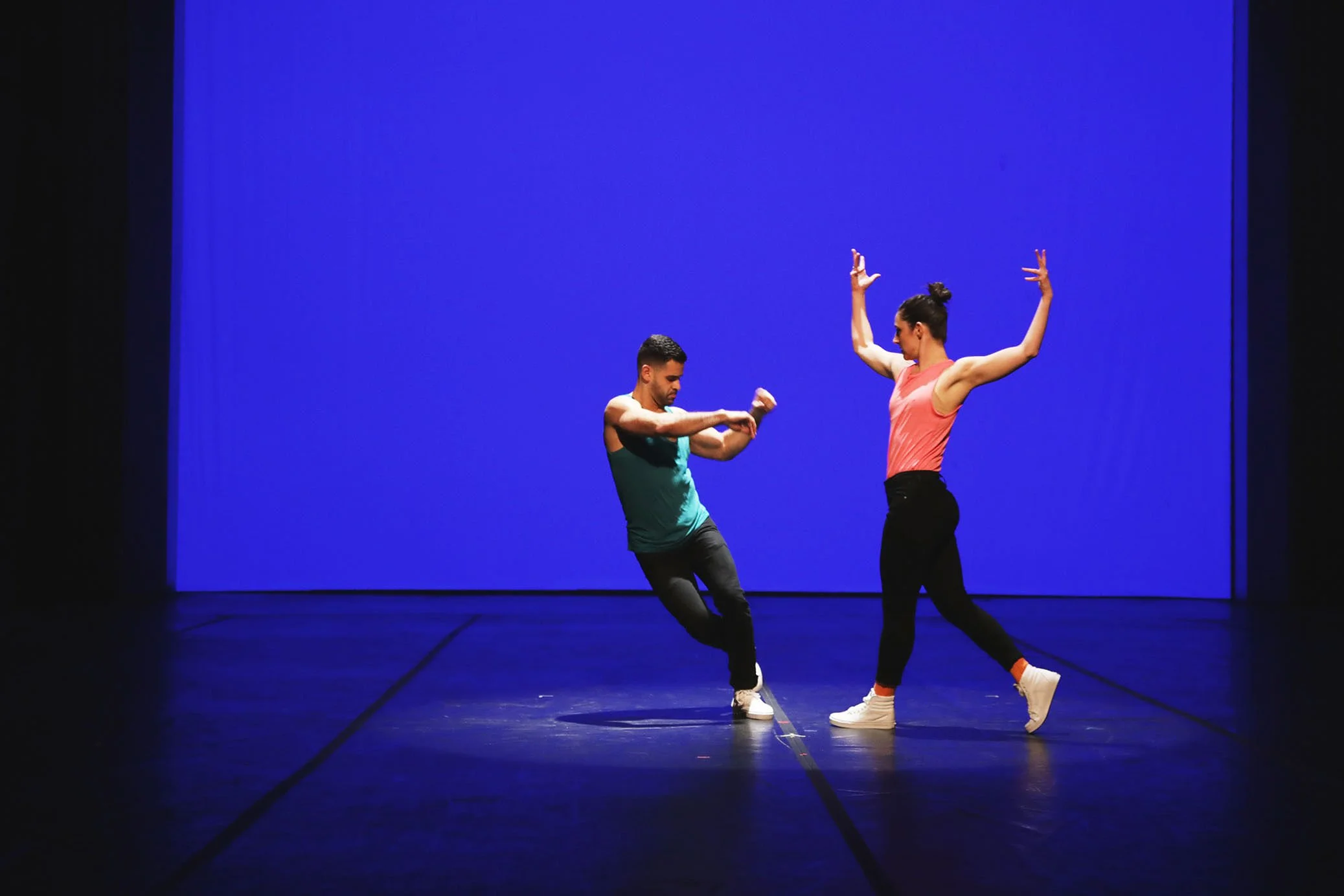Two dancers perform on a stage with a vibrant blue background, one male and one female, both wearing colorful workout clothes, striking expressive dance poses.