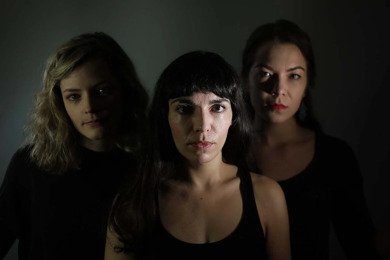 Three women posing in a dark setting with dramatic lighting, focusing on their faces.