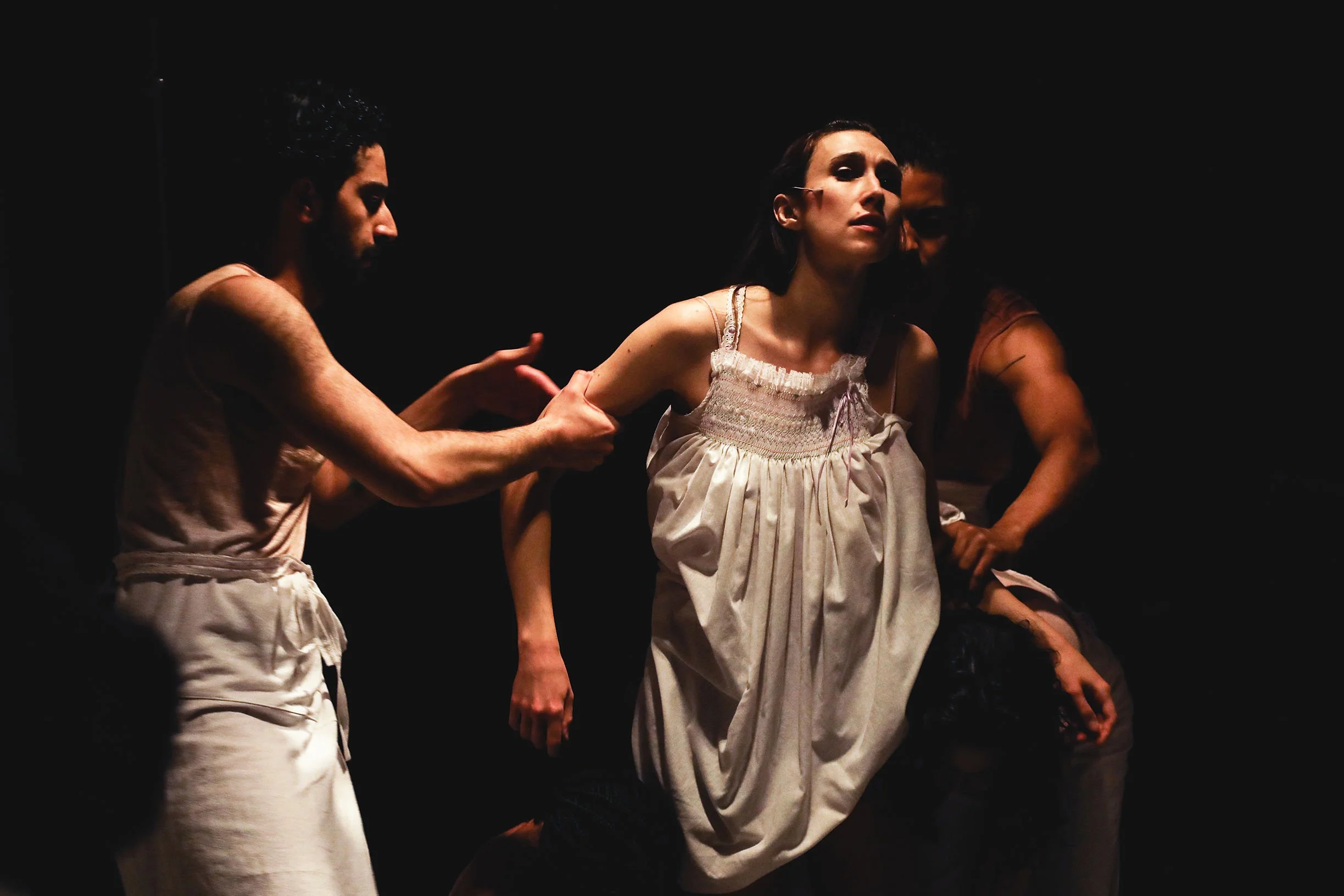 The image depicts several performers dressed in historical costumes, with some wearing cream-colored dresses and others wearing similar attire, engaged in a theatrical scene on a dark stage, indicating a dramatic performance.
