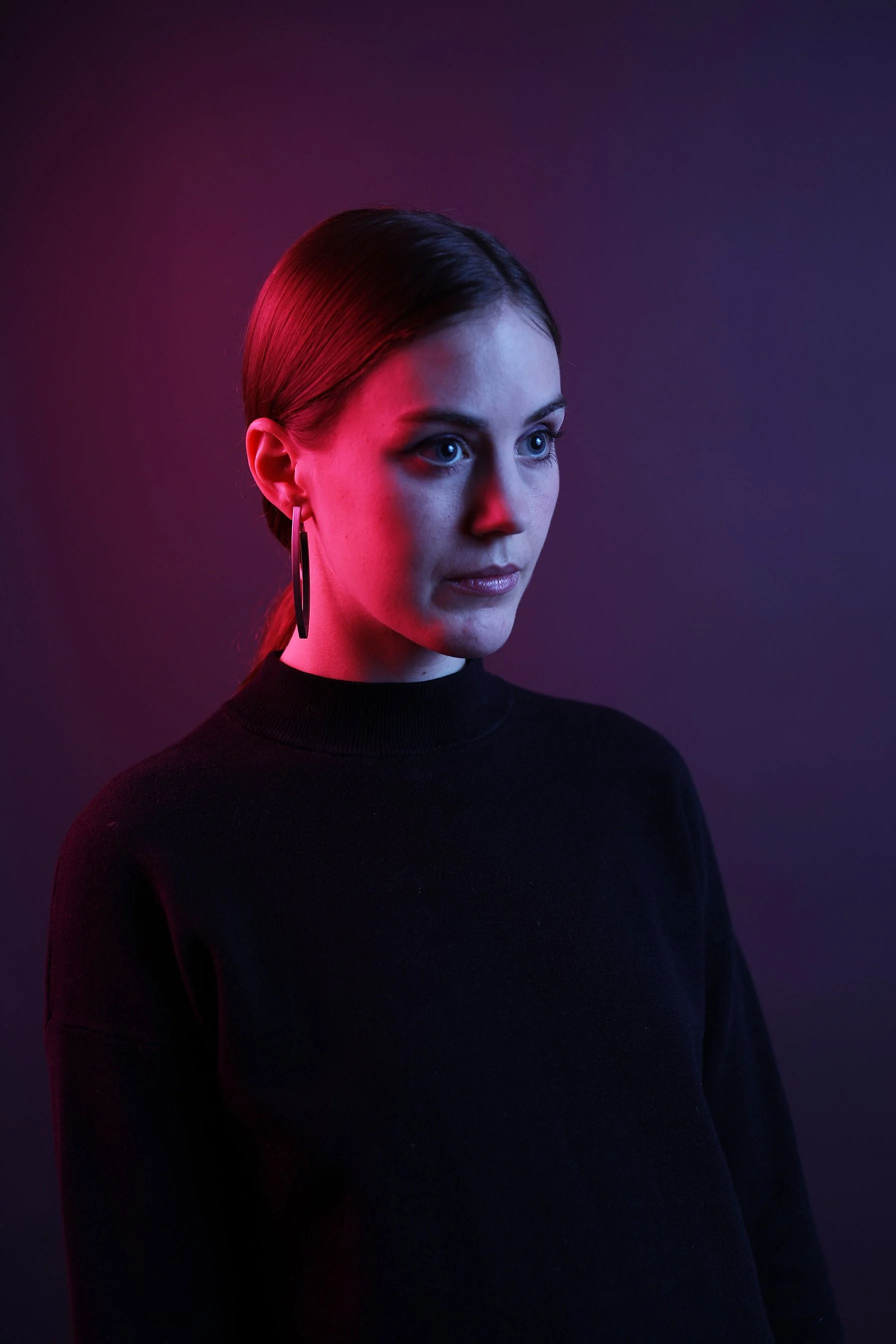 A woman with dark hair pulled back, wearing hoop earrings and a black turtleneck, illuminated with pink and purple lighting.