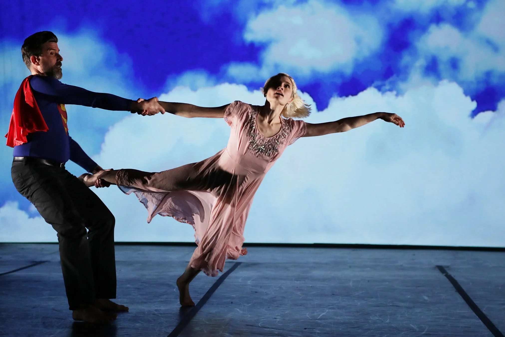 A male performer dressed as Superman holding a female ballet dancer in a pink costume, with both performing on a stage against a backdrop of a cloudy sky.