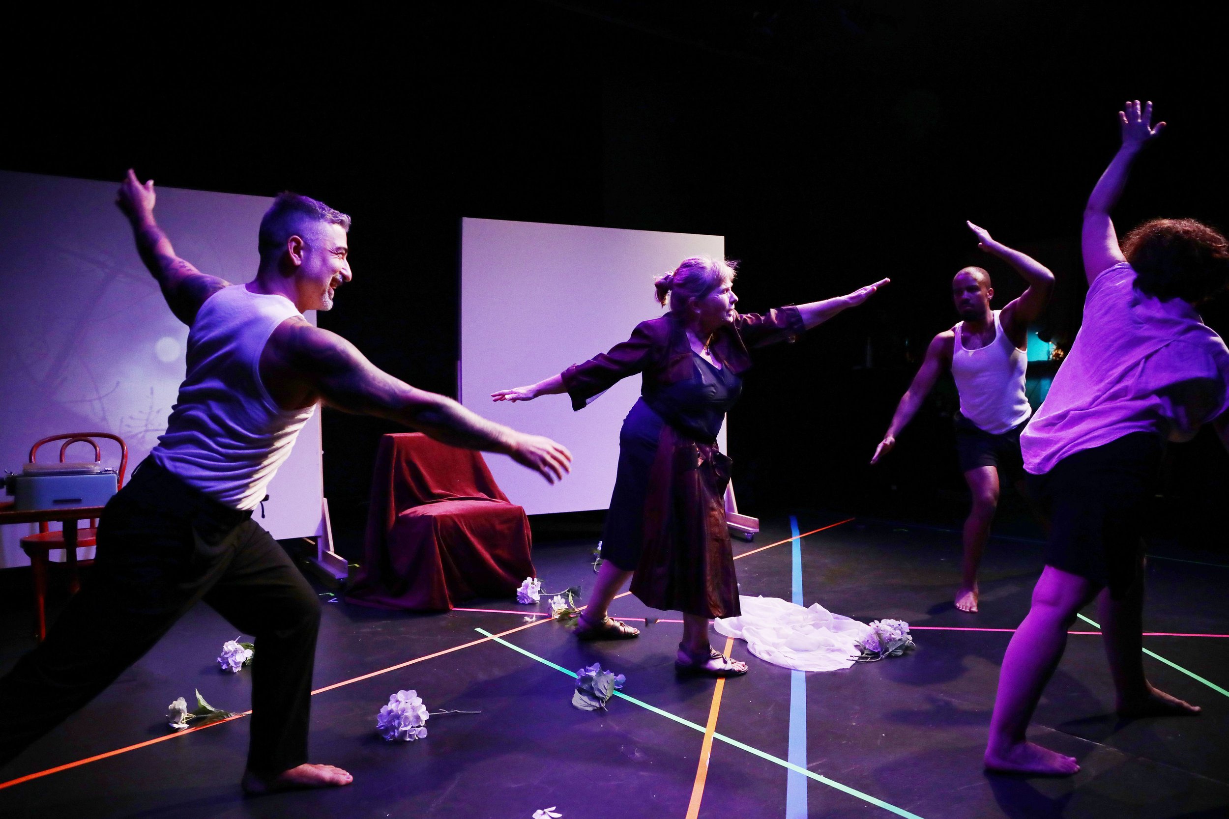 People engaging in a theatrical performance on stage, with some reaching out and others posed in expressive movements, surrounded by scattered flowers and minimal props, with black background and stage lighting.
