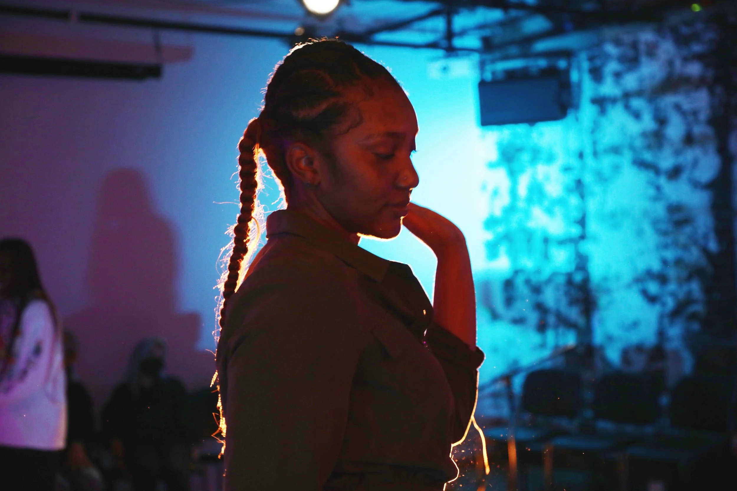 A woman with braided hair in a dimly lit room with blue and purple lighting, standing with her eyes closed and her hand near her face.