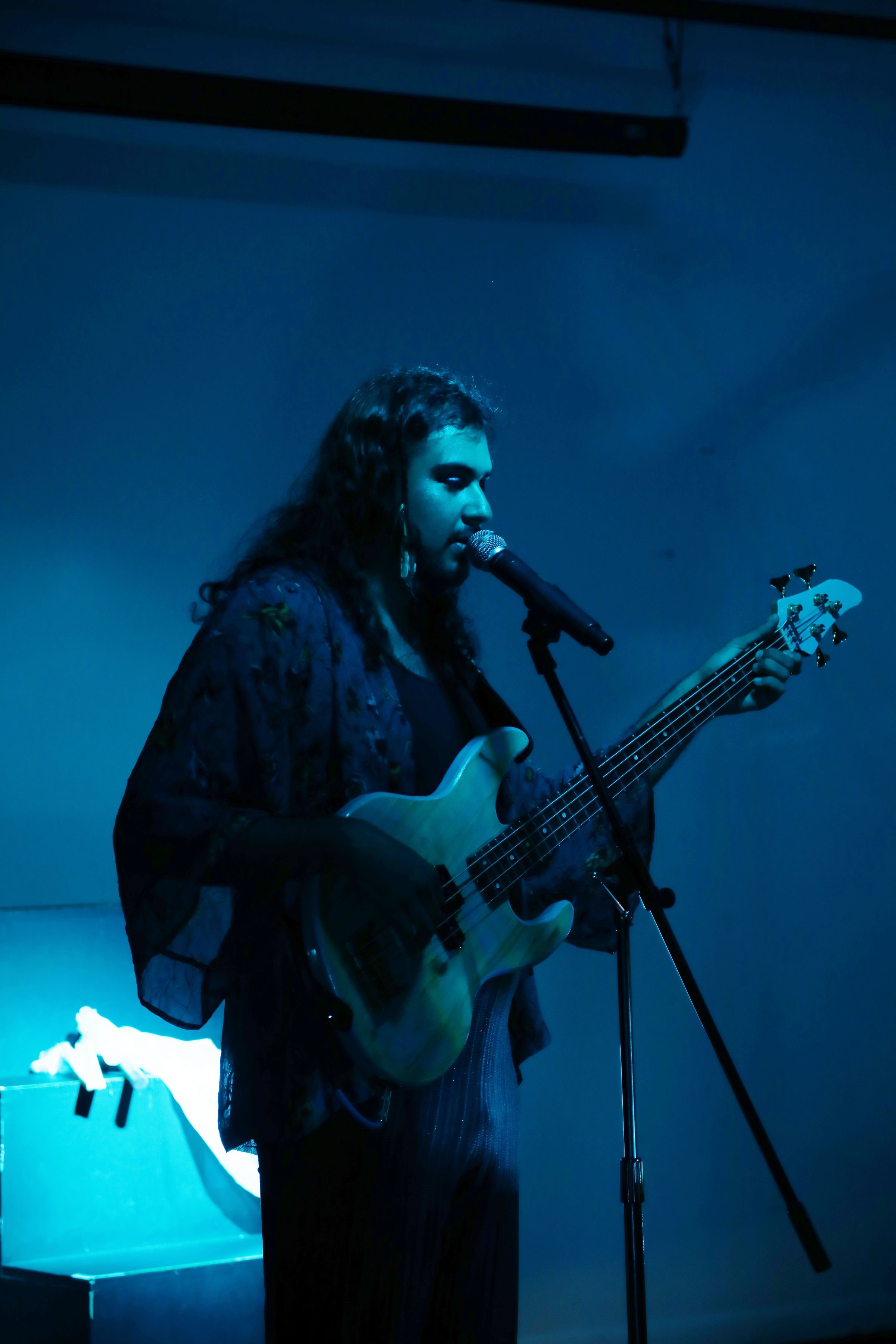 A woman with curly hair singing into a microphone while playing an acoustic guitar on a dimly lit stage during a performance.