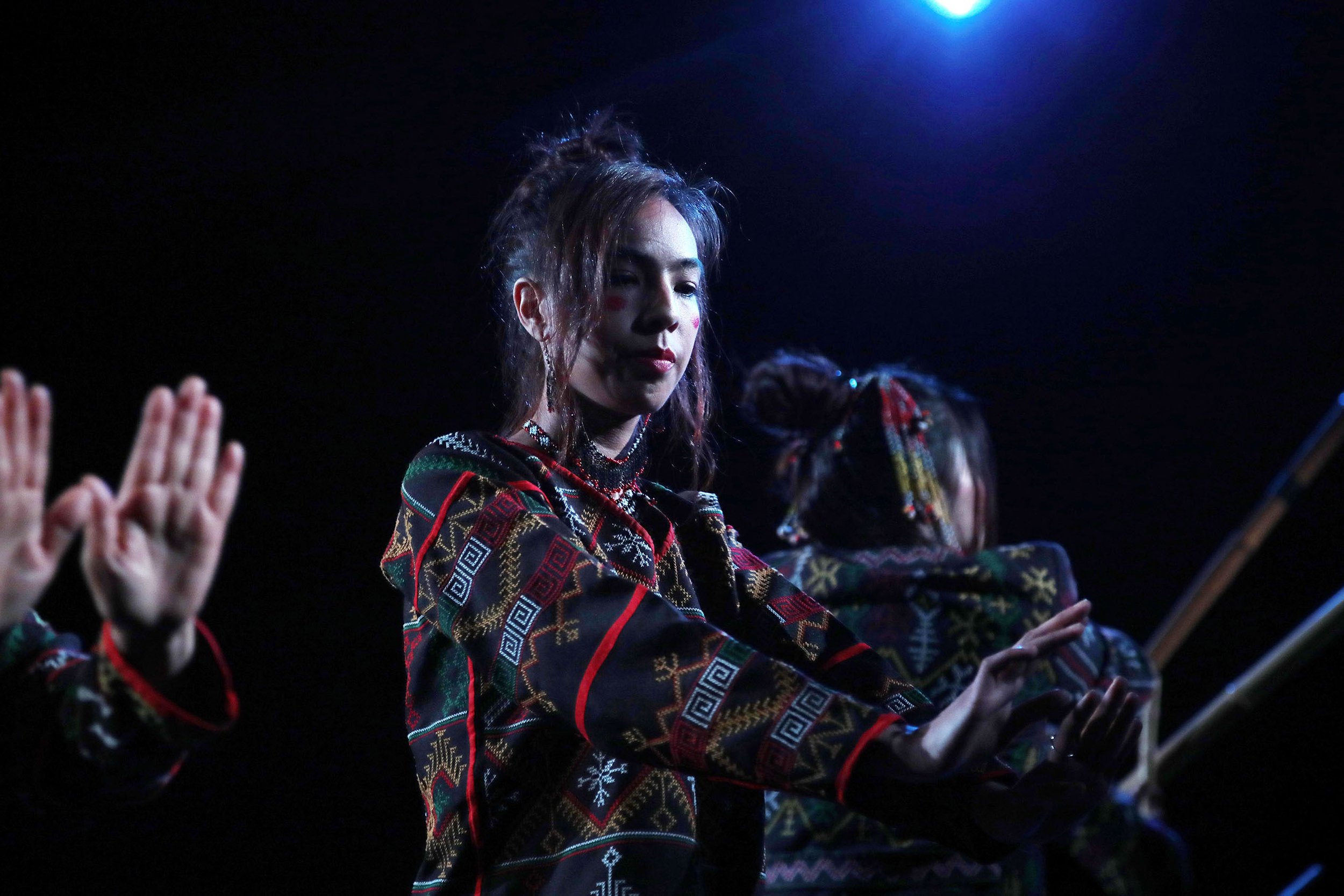 A young woman with long dark hair styled in a messy bun, wearing a colorful embroidered sweater with geometric patterns, is seen with her eyes closed and hands raised in a gesture of respect or celebration. The setting appears to be a stage or perfor