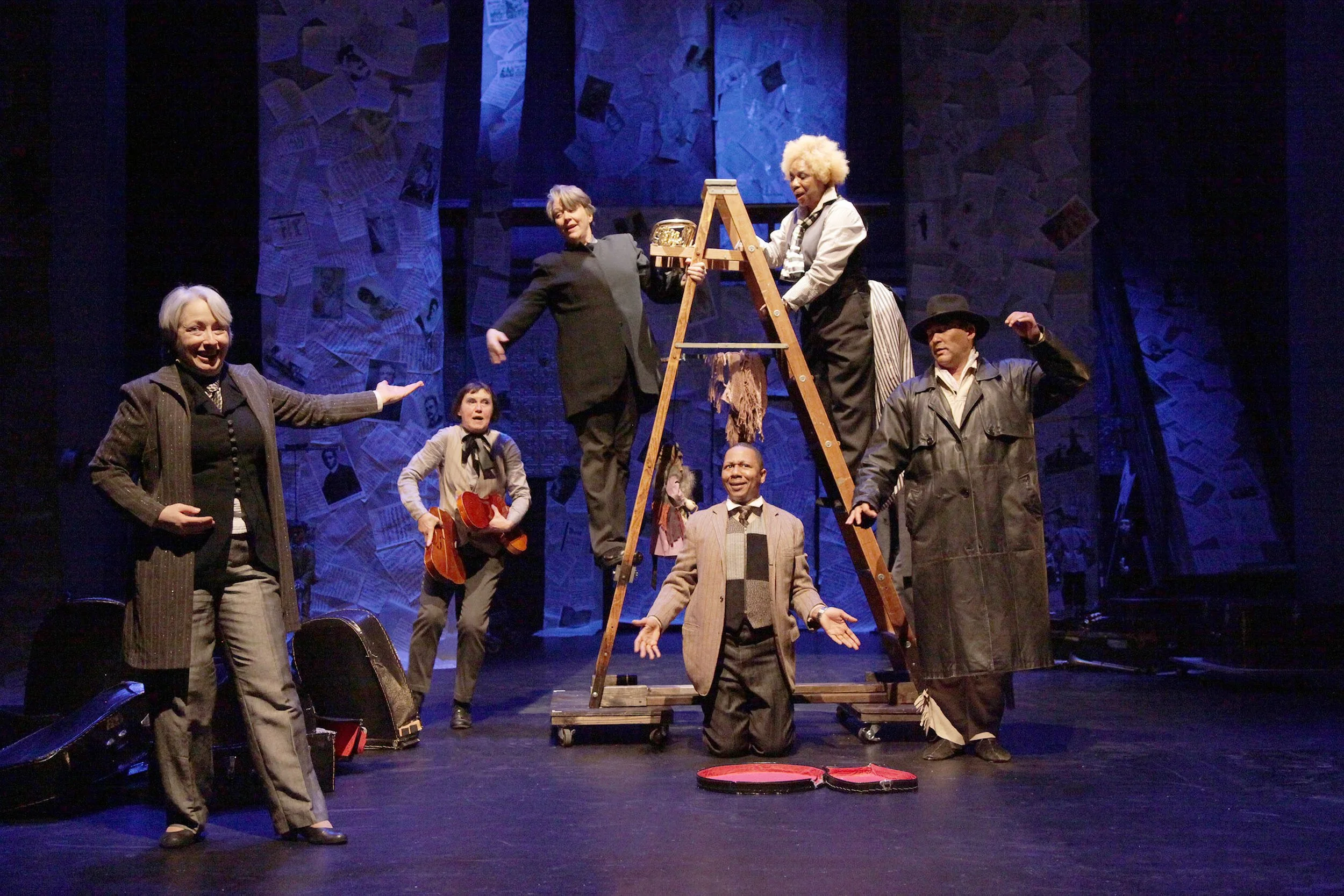 A theatrical stage scene with actors dressed in vintage costumes, including suits, coats, and hats. Some actors are standing, while others are on a wooden platform or ladder. The background features a textured blue wall decorated with newspapers.