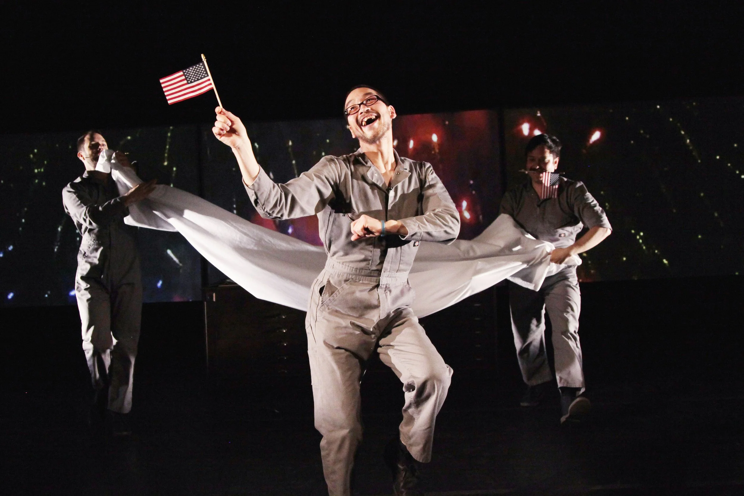 A group of performers dancing on stage, with the central person holding an American flag, smiling and laughing, with a dark background and starry pattern.