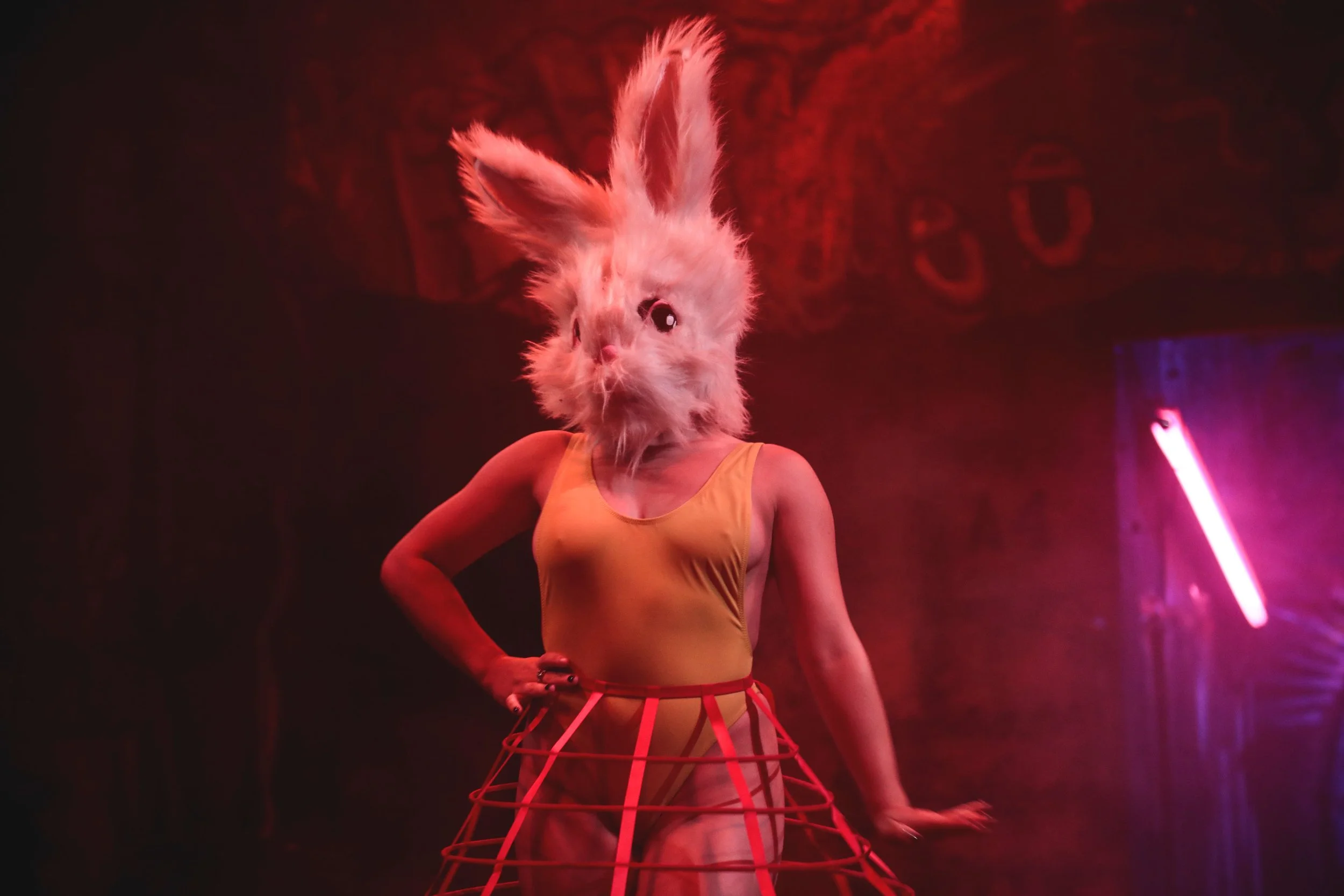Person wearing a white furry rabbit mask, orange tank top, and orange skirt with wireframe design, standing in a dimly lit room with pink and red lighting effects.
