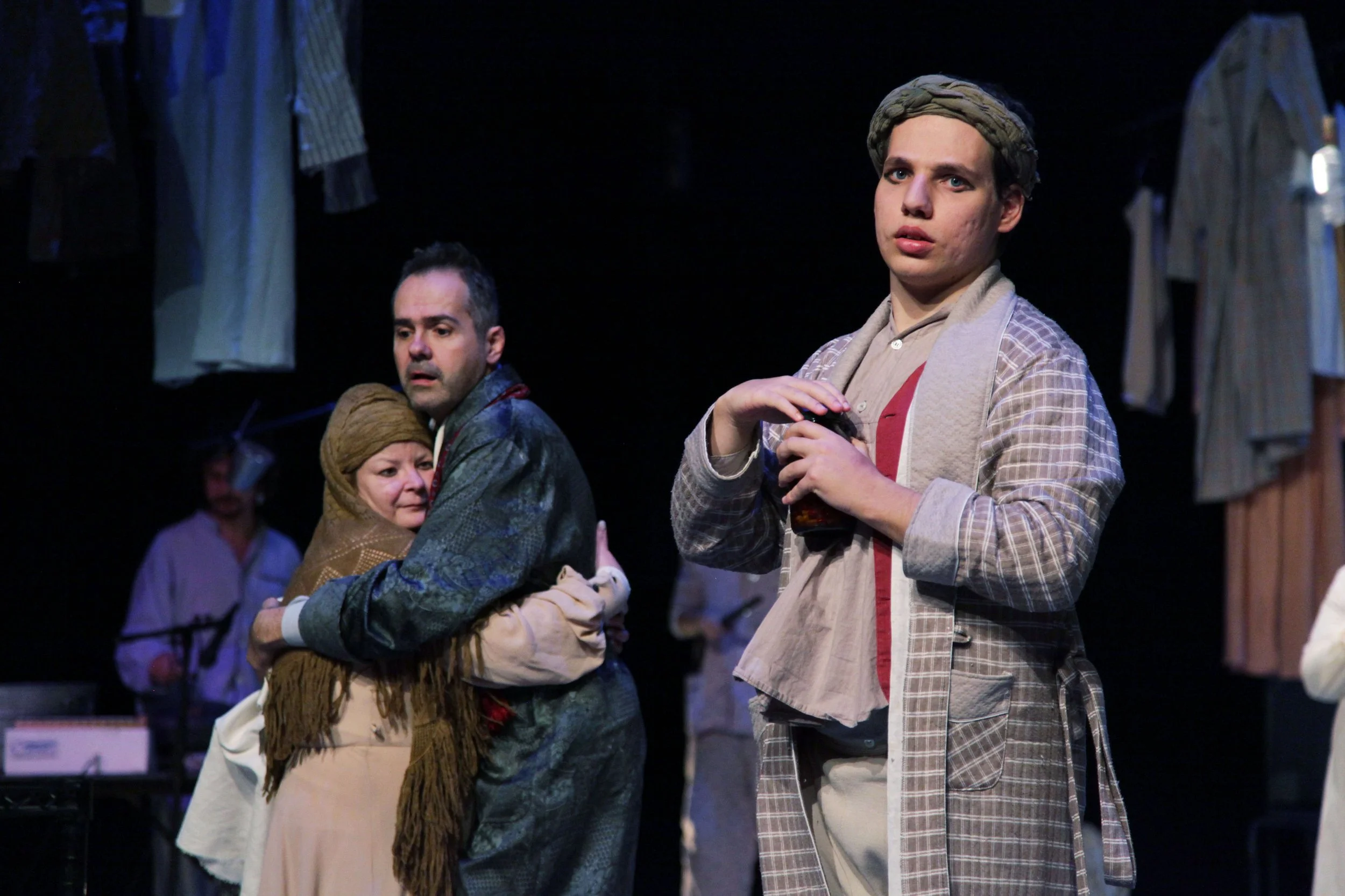 Four people standing in a dark setting, with hanging clothes visible. The central figure is a young woman holding a drink, wearing a plaid jacket and headscarf. To her left, an older woman and man look on, and another woman is partly visible in the b