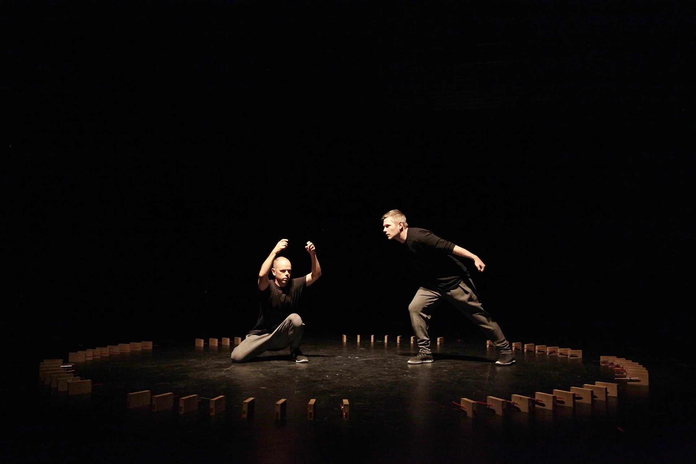 Two male performers on a dark stage, one kneeling and the other standing, engaged in a dance or theatrical performance, with wooden blocks arranged on the stage floor.