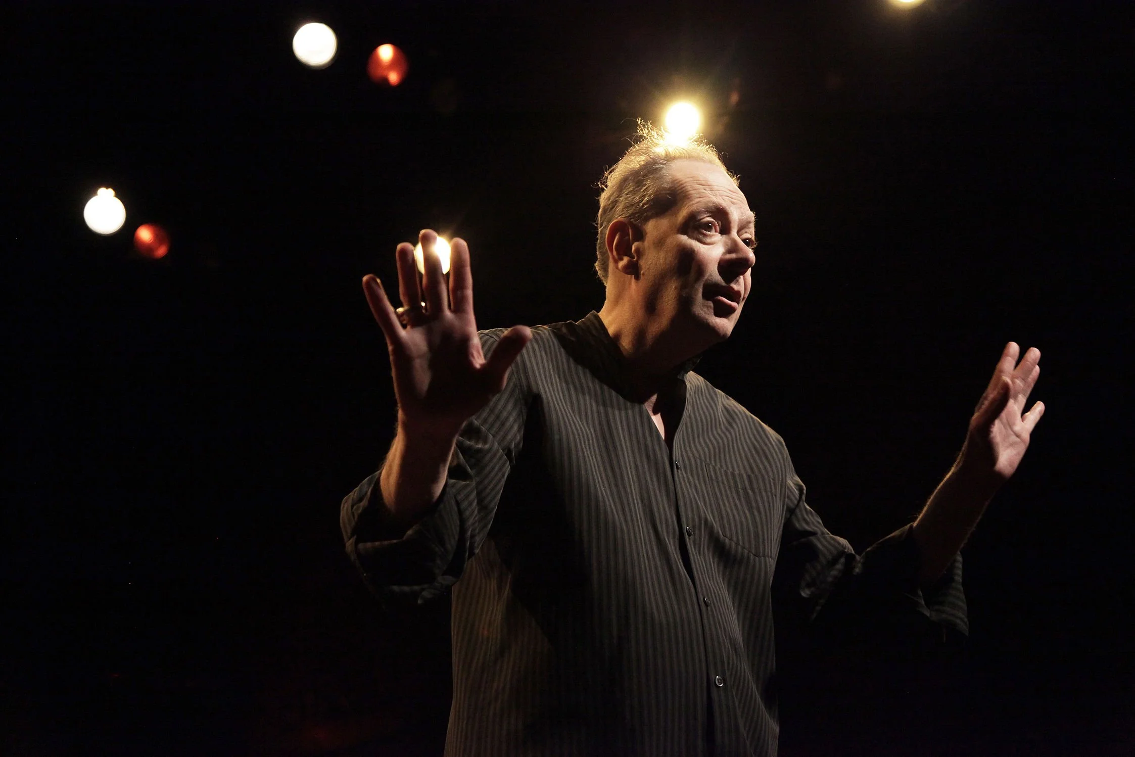 A man with gray hair and a striped shirt speaking or performing on stage, with stage lights illuminating his head and hands raised in front of him.