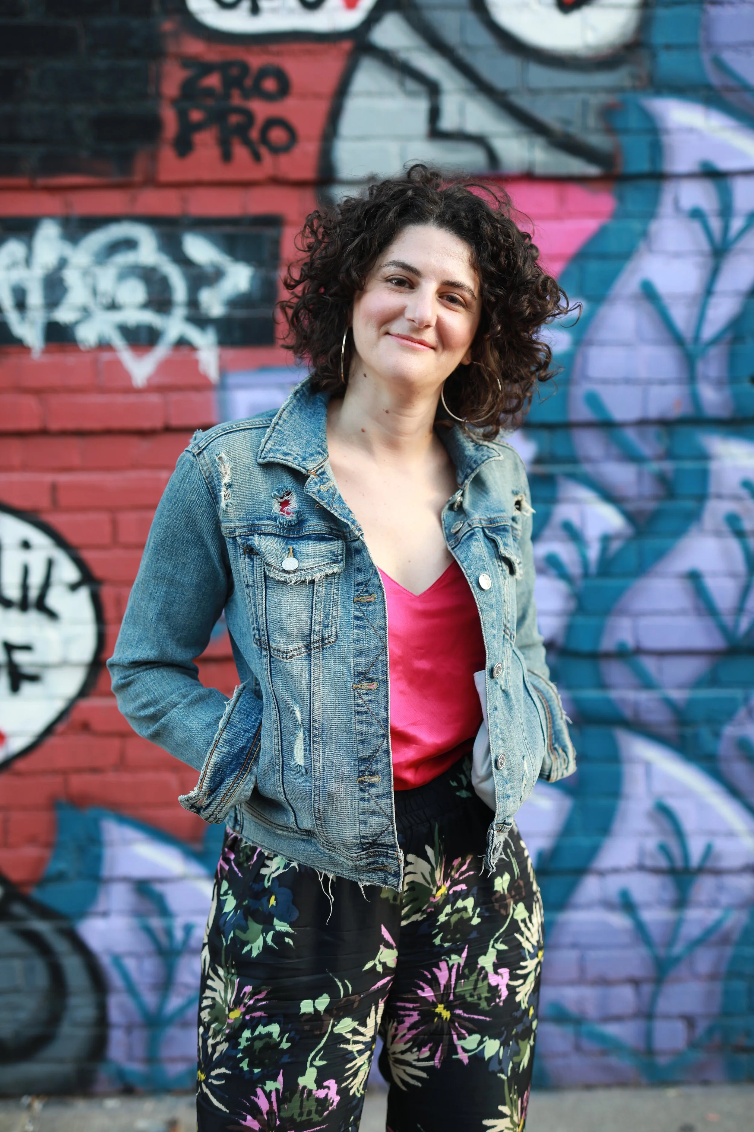 A young woman with curly dark hair wearing a denim jacket, pink top, and floral pants standing in front of a graffiti wall with a smile.