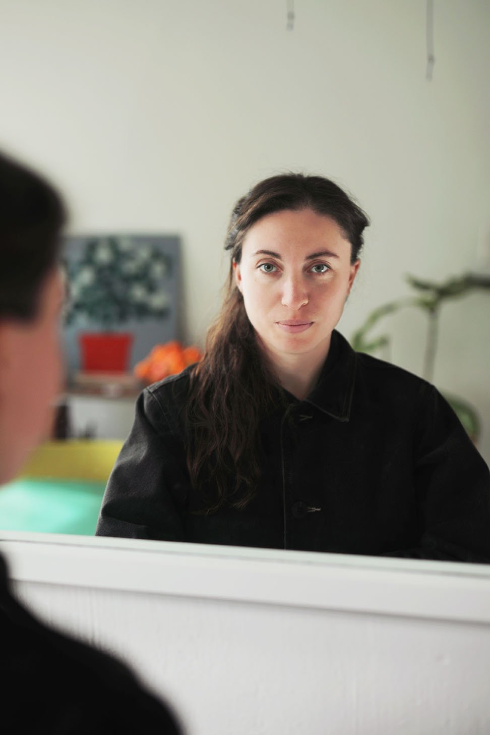 A young woman with long dark hair wearing a black jacket, gazing intently at a person whose back is visible in the foreground.