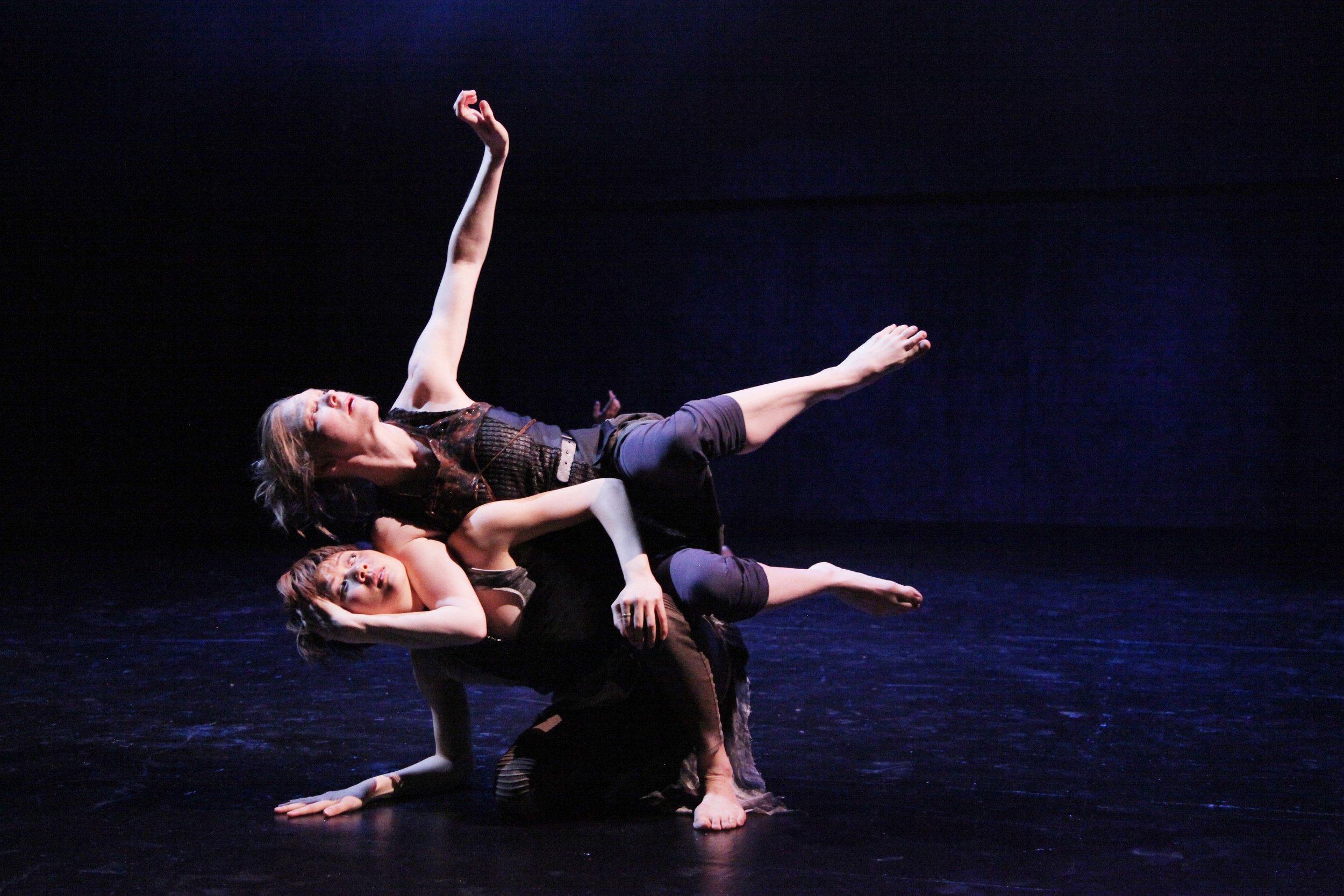 Dancer performing an acrobatic pose on stage with a dark background, two dancers are supporting her from below.