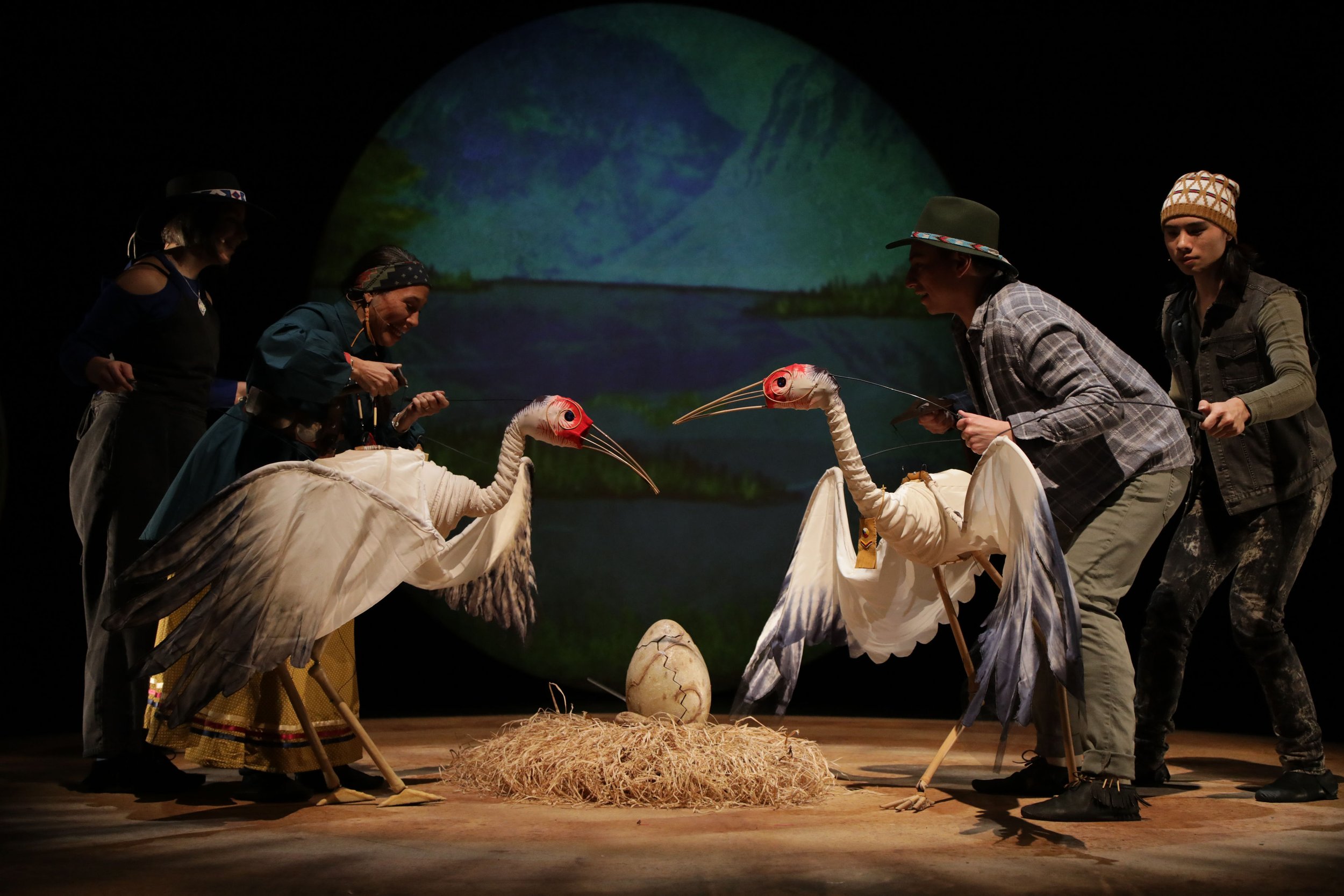 People dressed in costumes performing a scene with puppet birds, a large nest, and an eggshell on stage, with a painted mountain landscape in the background.
