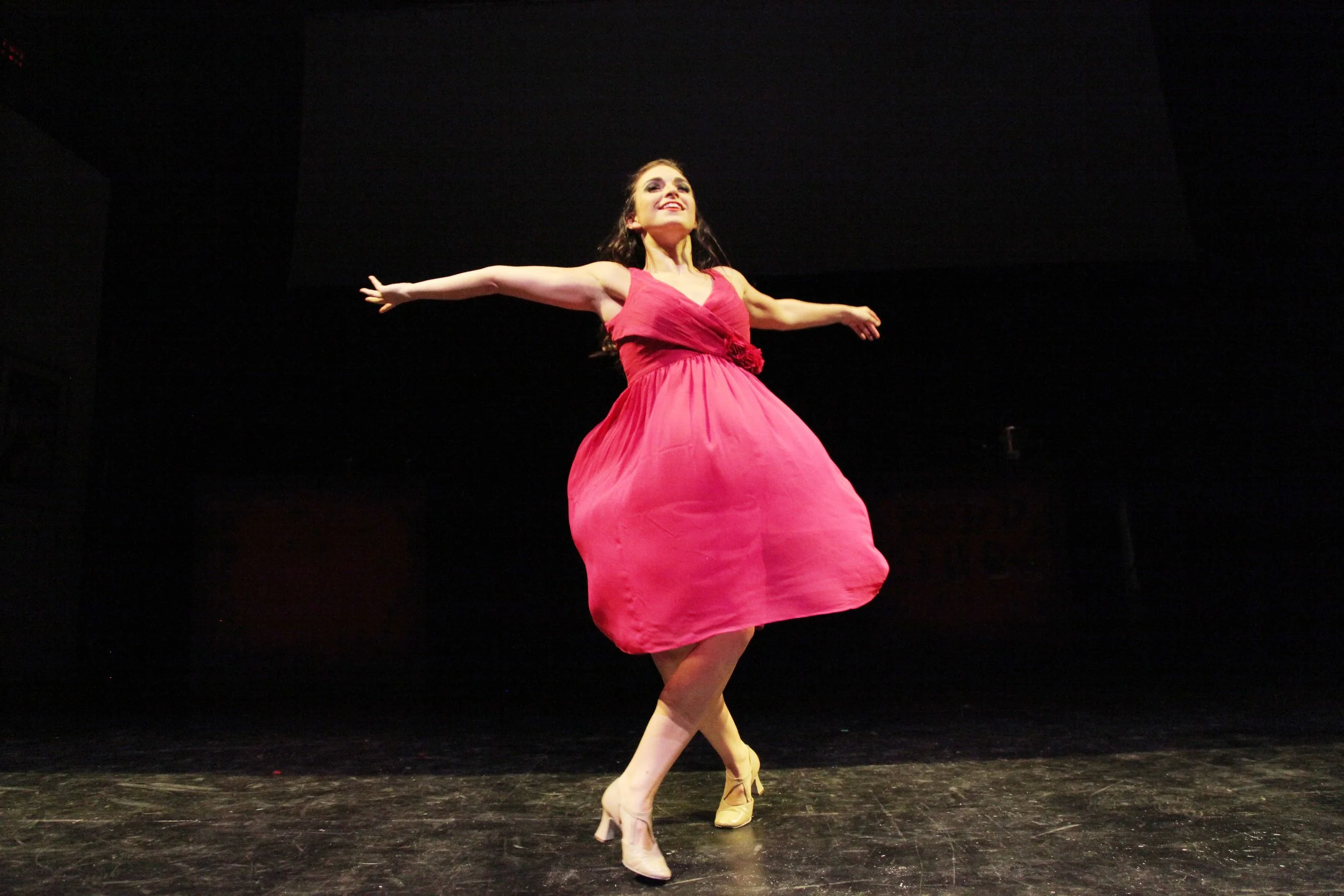 A woman dancing on stage in a pink dress with arms outstretched, dark background, and stage floor visible.
