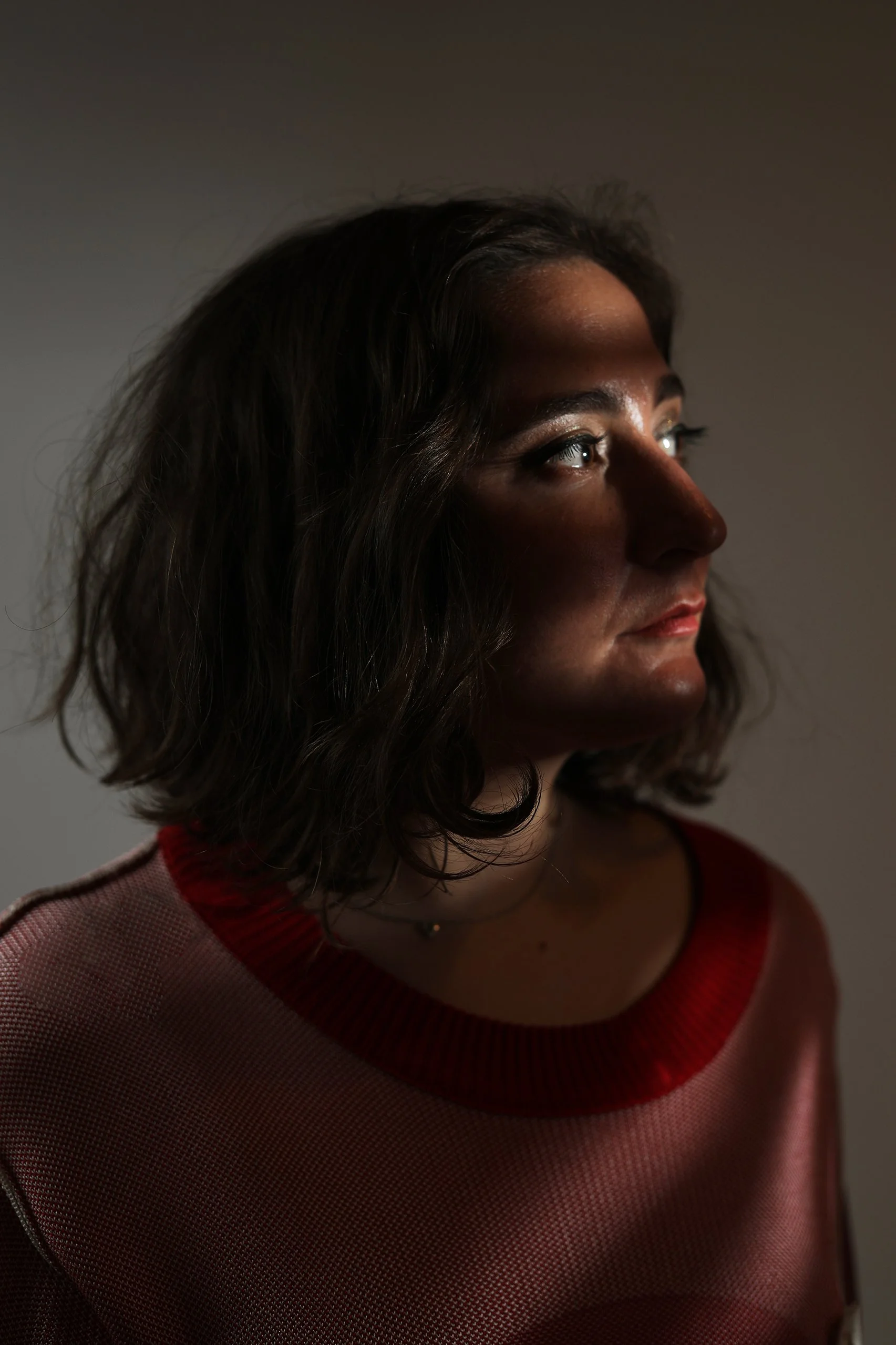 A young woman with dark, wavy hair looking to the right, wearing a red and beige sweater in a dimly-lit setting.