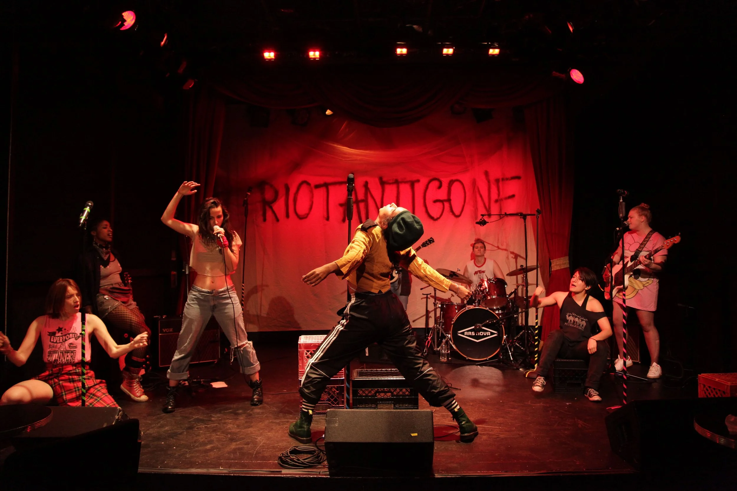 A lively stage performance featuring multiple young performers, including a woman singing into a microphone, a person dancing with a hat and striped socks, and another playing the guitar. The backdrop reads 'RIOTANTIGONE' with red stage lighting.