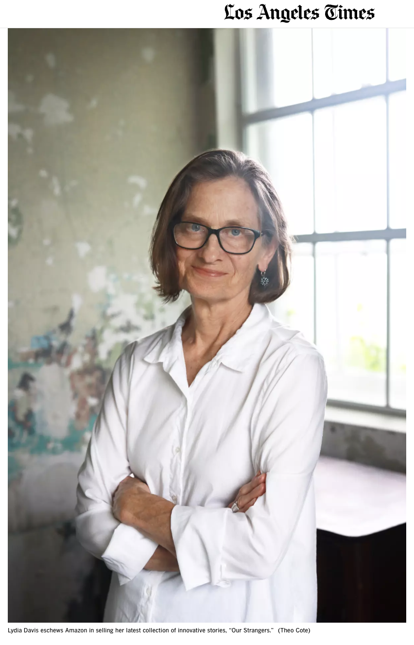 A woman with shoulder-length brown hair, wearing glasses and a white button-up shirt, standing with her arms crossed in front of a window with natural light, in a room with a partially painted or distressed wall.