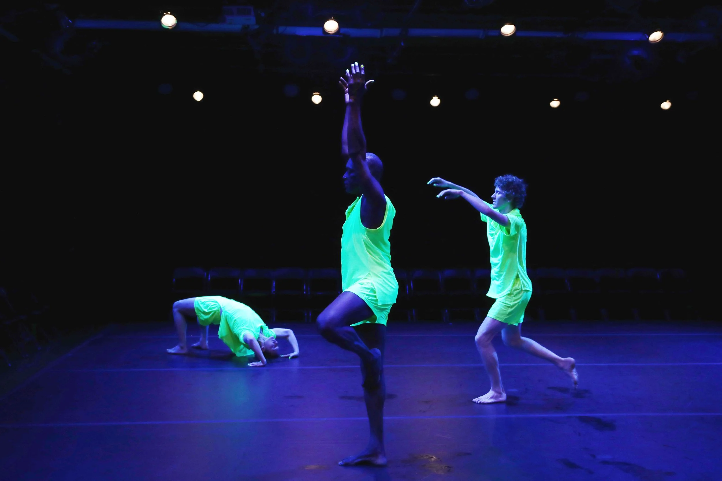 Dance performance featuring two dancers in neon yellow costumes on a dark stage, one standing with arms raised and the other in a plank position.