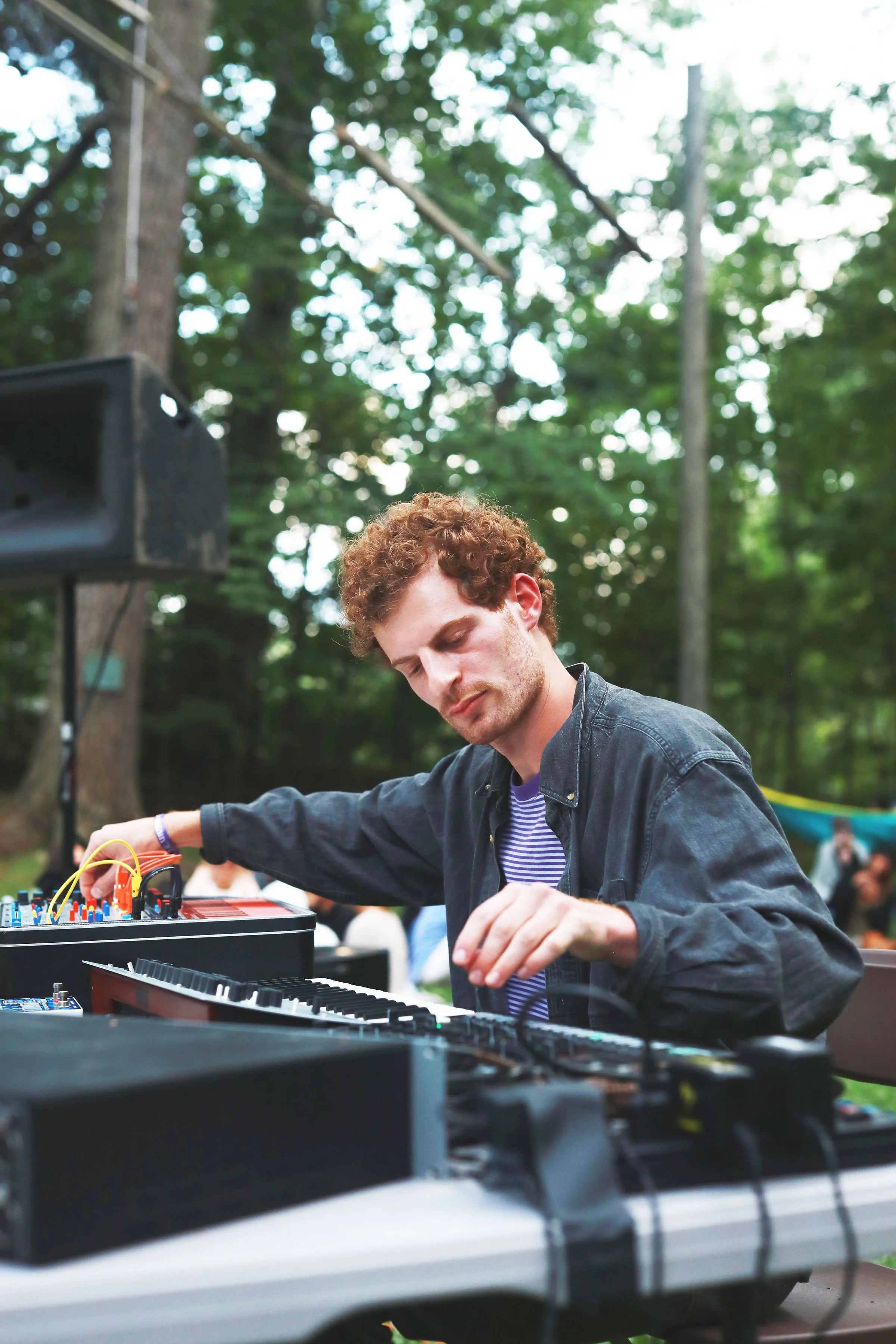 A young man with curly red hair and a striped shirt is performing electronic music with a synthesizer and control board outdoors in a wooded area, with trees and blurred people in the background.