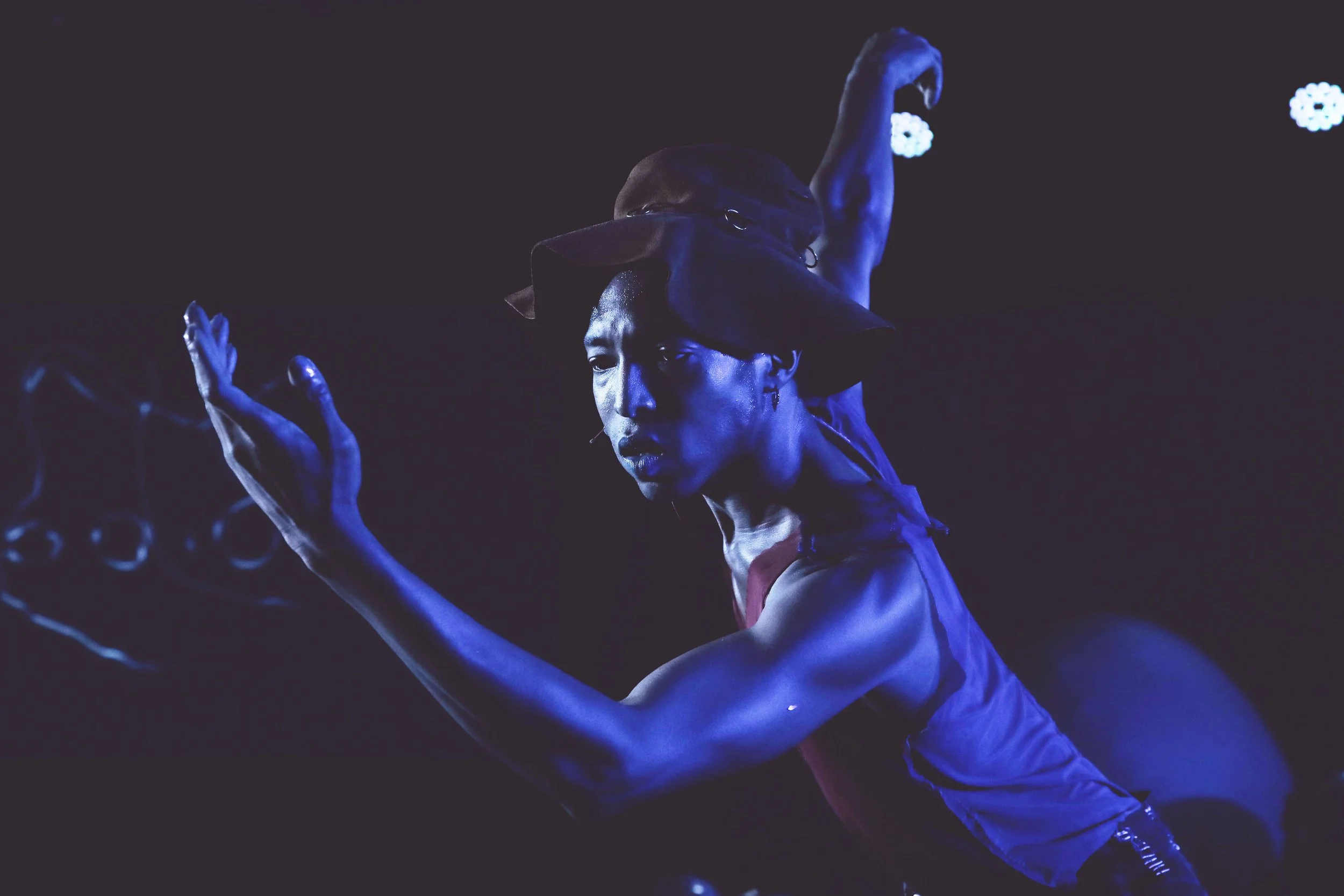 A performer with dark hair in a ponytail, dressed in a sleeveless outfit, poses with one arm raised and the other extended, illuminated by dramatic blue lighting in a dark setting.