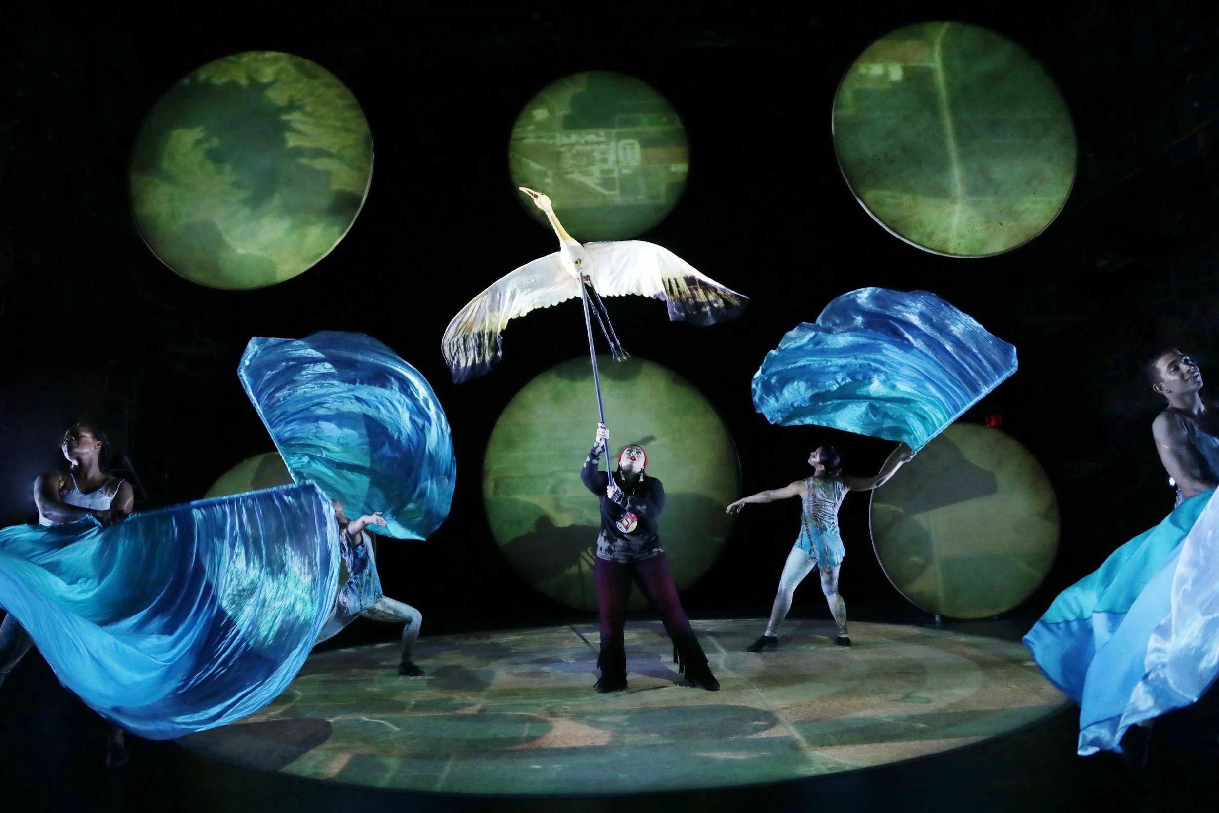 Performers in an artistic dance or circus act on stage with large blue fabric props and a woman holding a white bird, surrounded by green circular screens in the background.