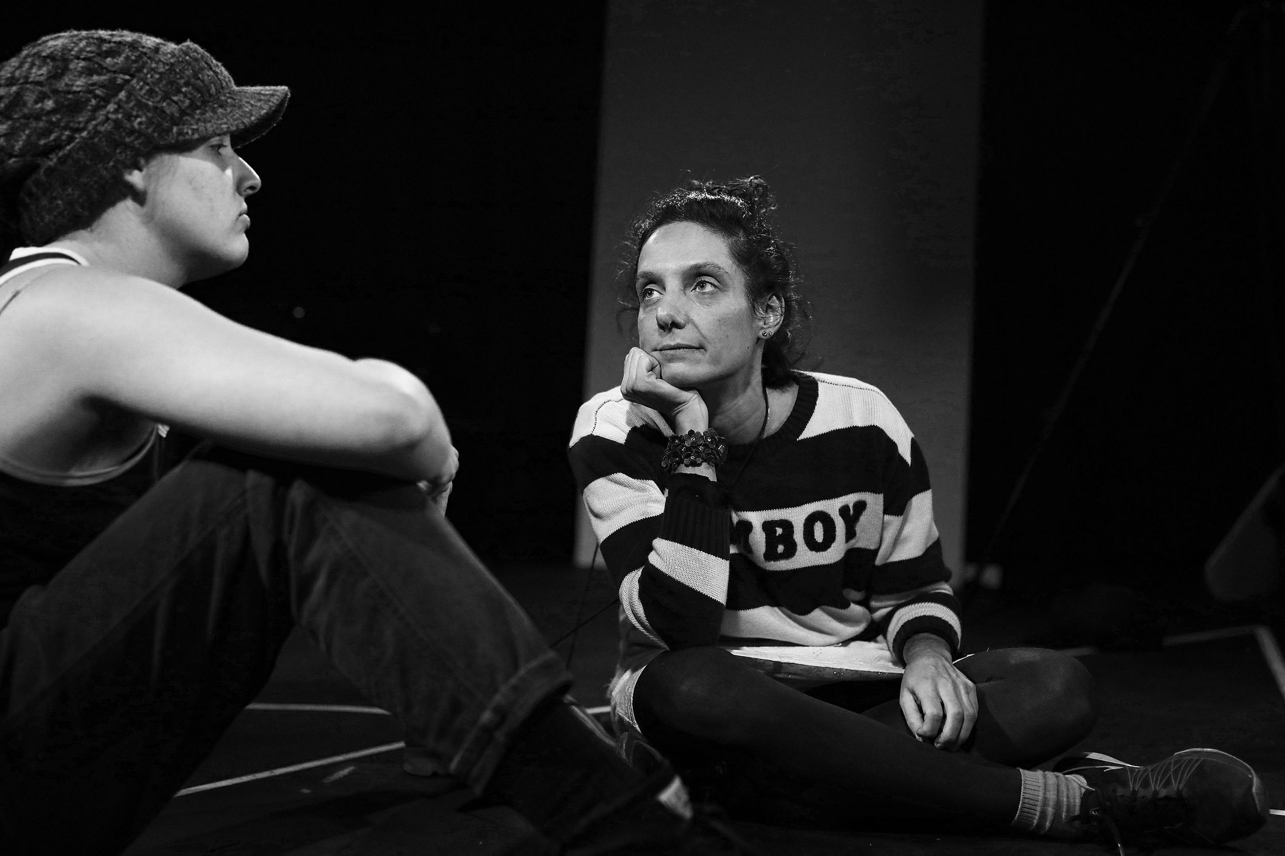 A woman with curly hair and striped sweater sitting on the floor, talking to a young person wearing a cap, both in a dark room.