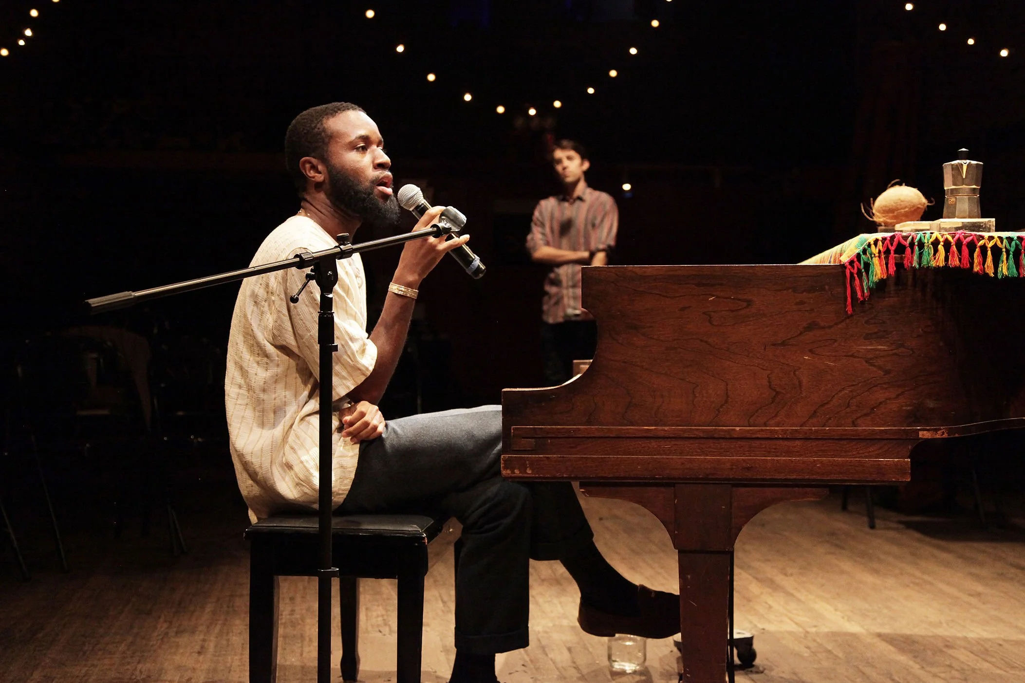 A man with a beard and dark skin sits at a piano, singing into a microphone in a dimly lit room. A woman with light skin, short hair, and a striped shirt stands in the background with arms crossed, watching him. The piano has a colorful tassel decora
