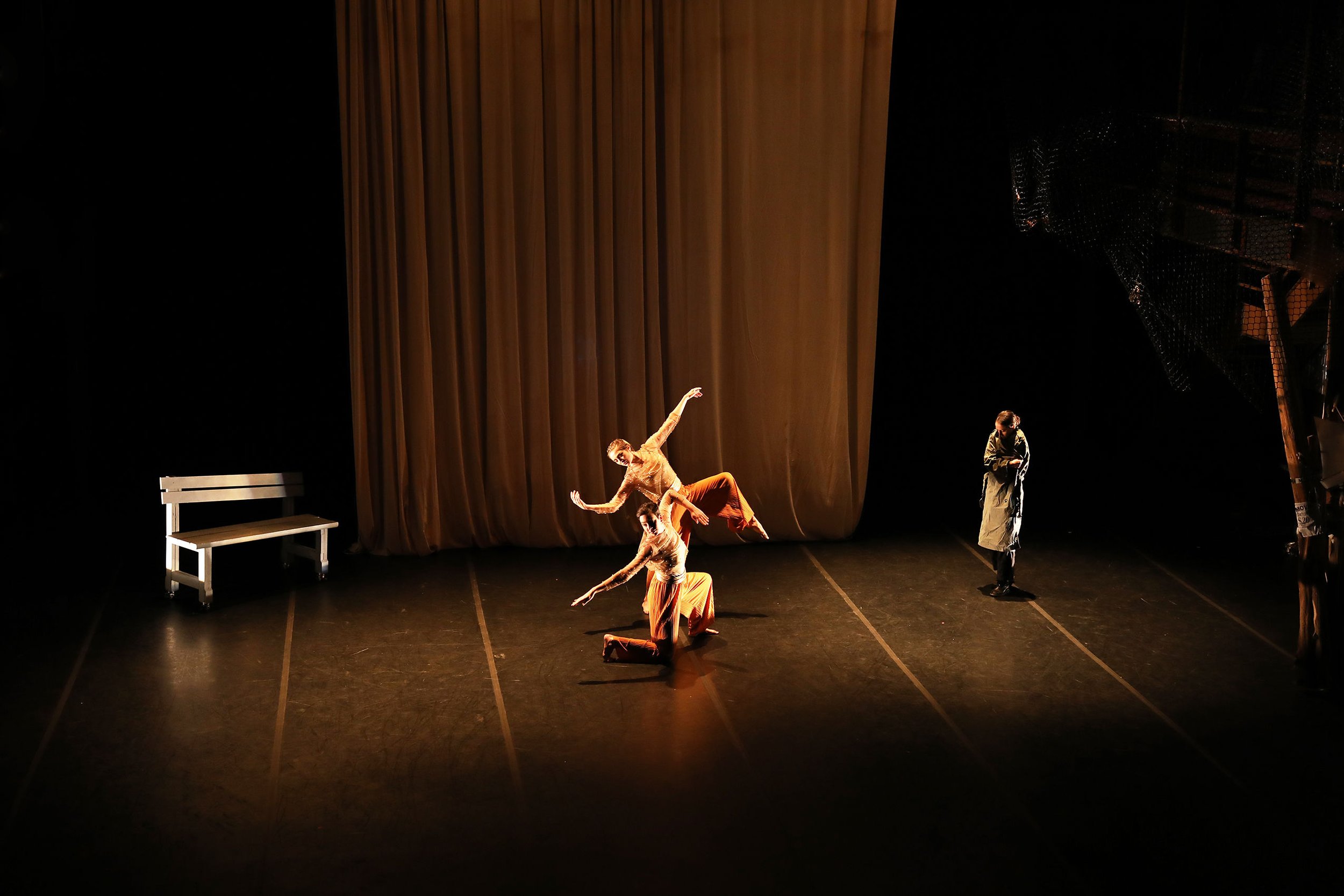 Dance performance on stage with four dancers, one standing, three in poses, a white bench on the left, a large curtain in the background, and stage lighting.