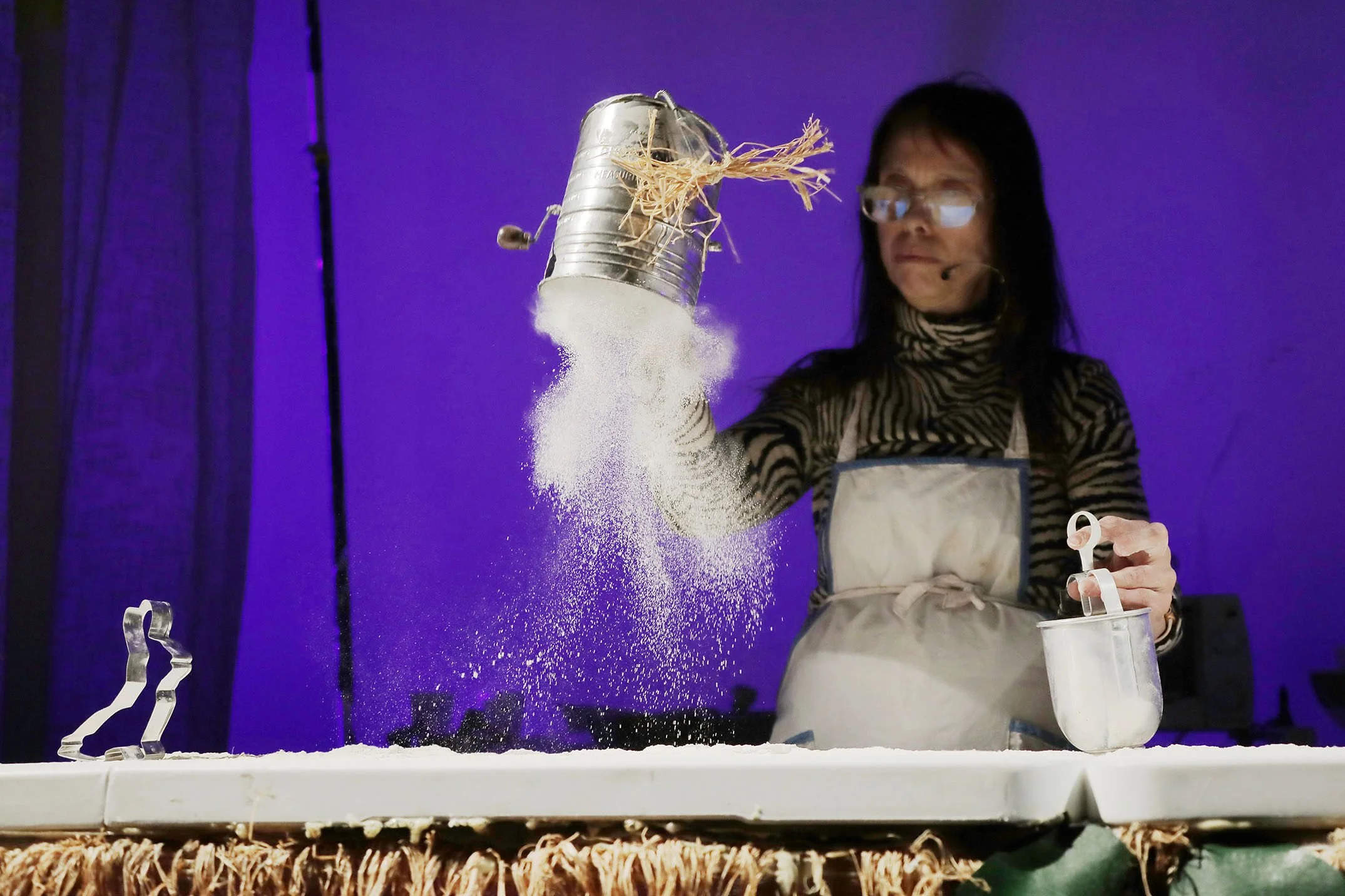 A woman wearing glasses and a striped shirt demonstrates a science experiment involving a tin can, straw, and white powder, with a purple background.