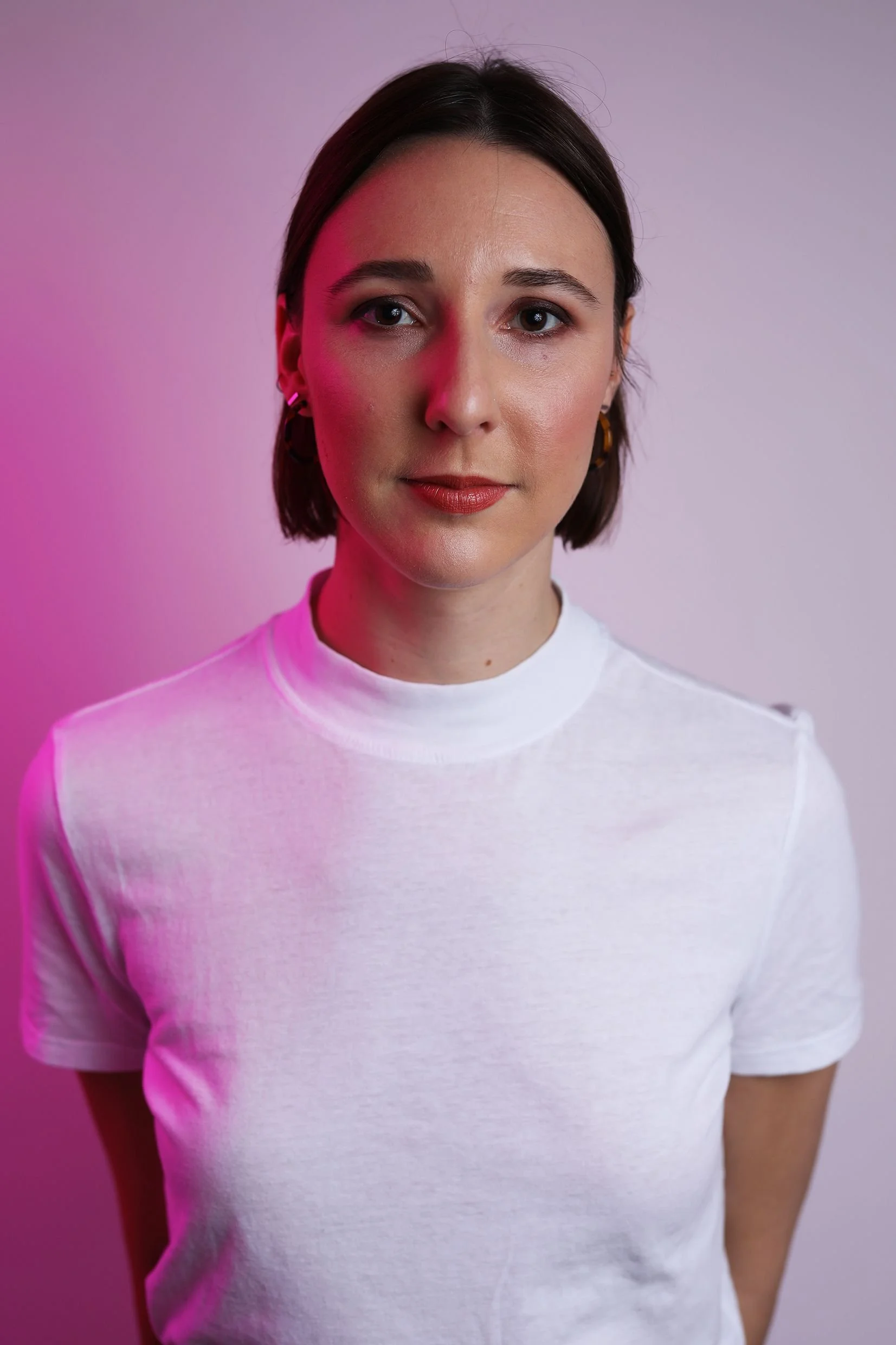 A young woman with short brown hair, wearing a white top, looking directly at the camera, with pink and purple lighting in the background.