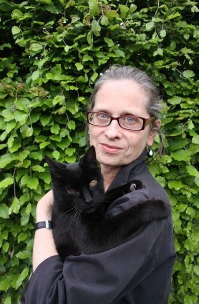 Woman with glasses holding a black cat in front of a leafy green background.