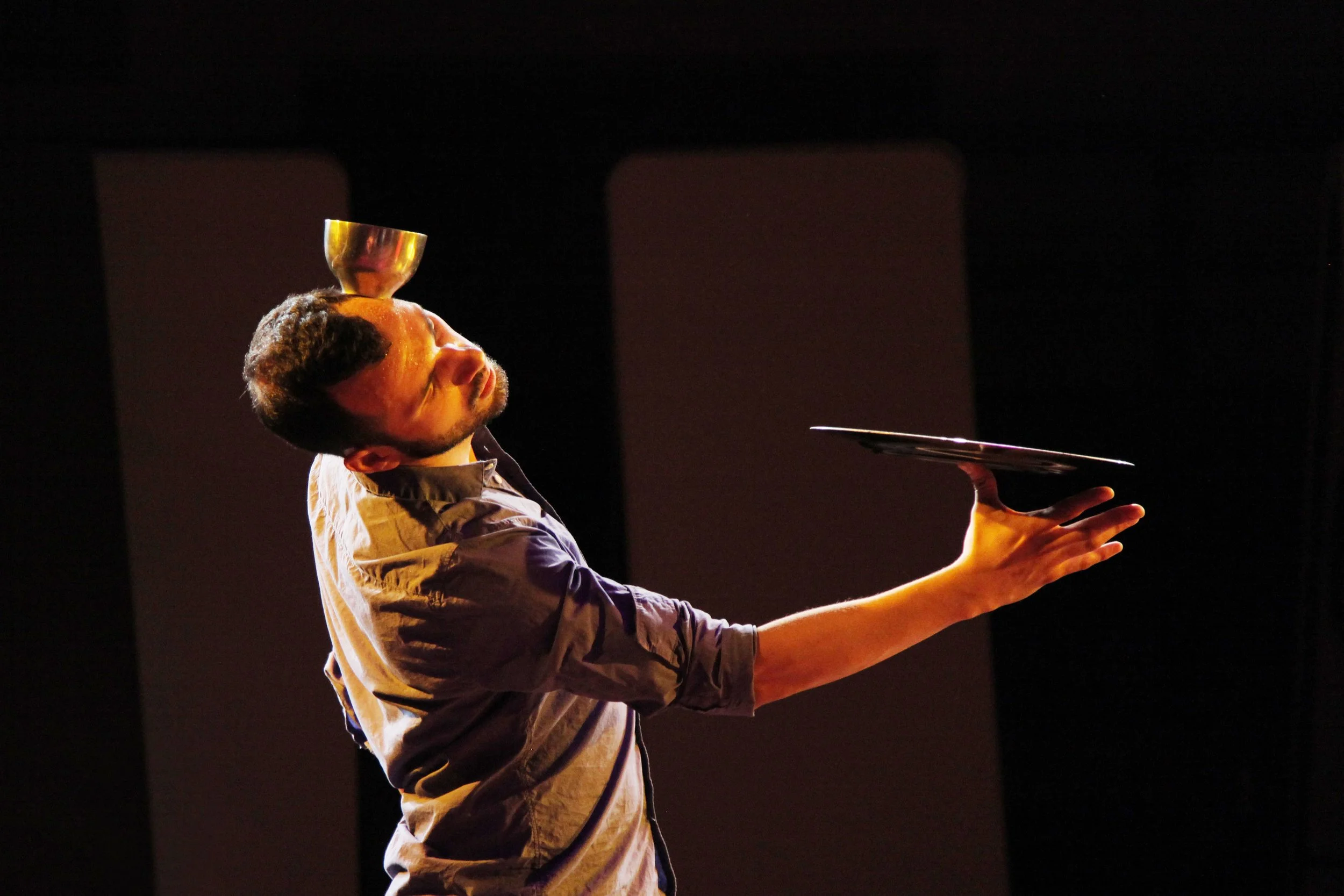 A man curves his body while balancing a metal bowl on his forehead and holding a tray with his hands in a dark room.