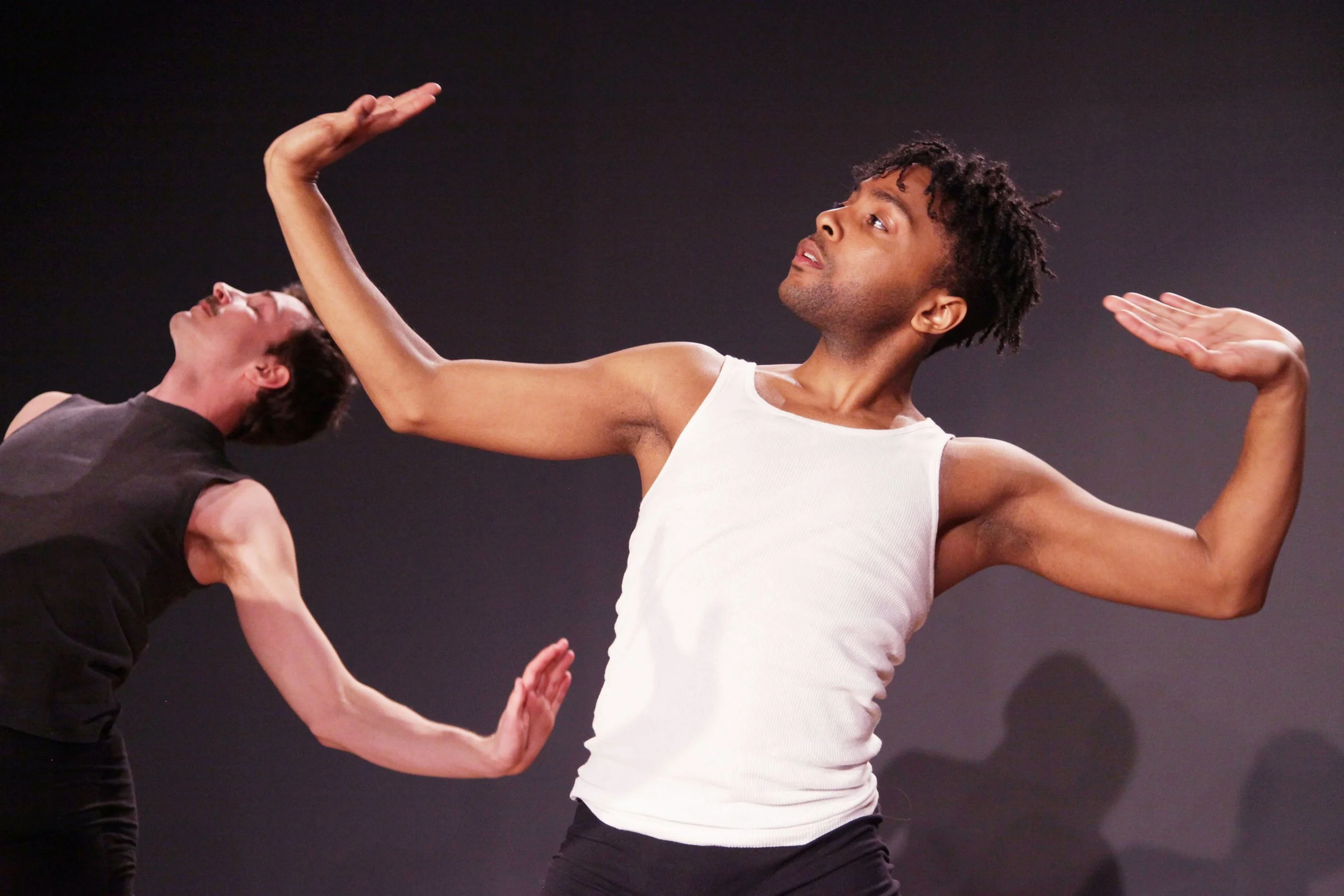 A dancer wearing a white tank top and black pants performs expressive movements with arms raised, while another dancer in black executes a pose with head tilted back. The background is dark.