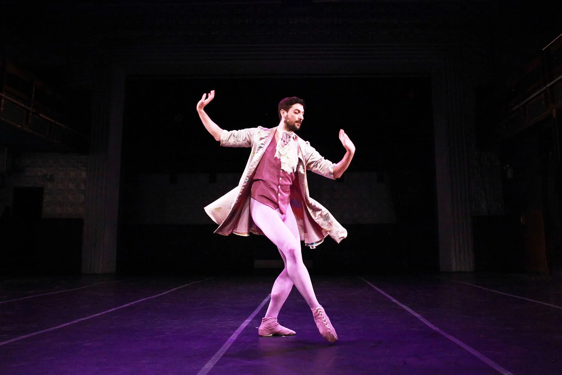Ballet dancer performing on stage in pastel-colored costume, standing on one foot with arms raised.