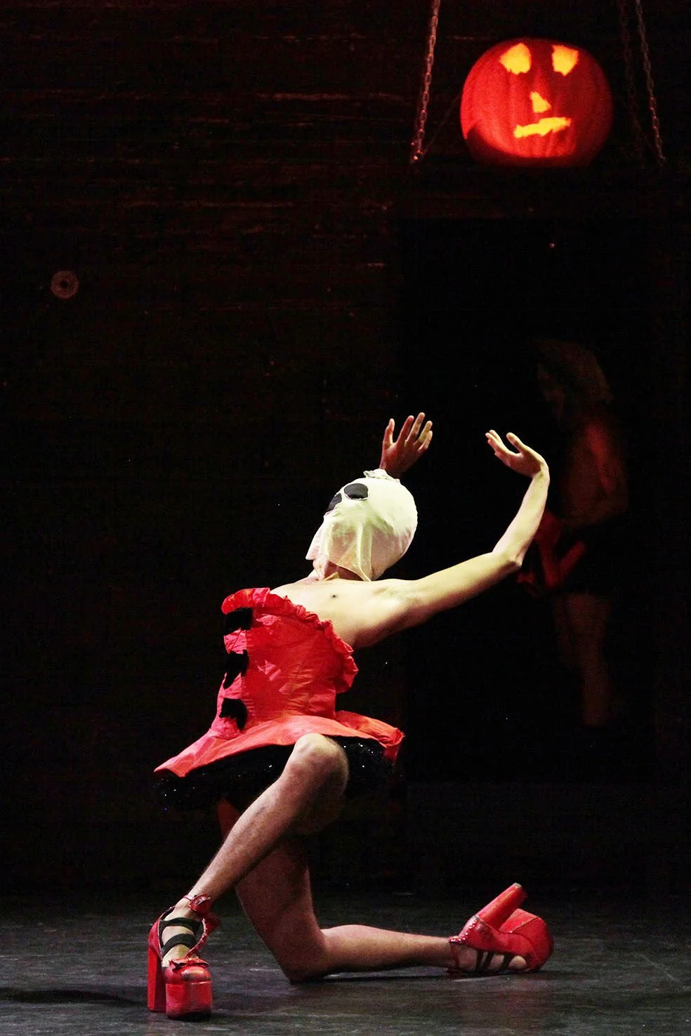 Performer wearing a mask resembling a skull, red dress, and red platform shoes, kneeling on stage with arms raised, halloween pumpkin lantern hanging above in a dark setting.