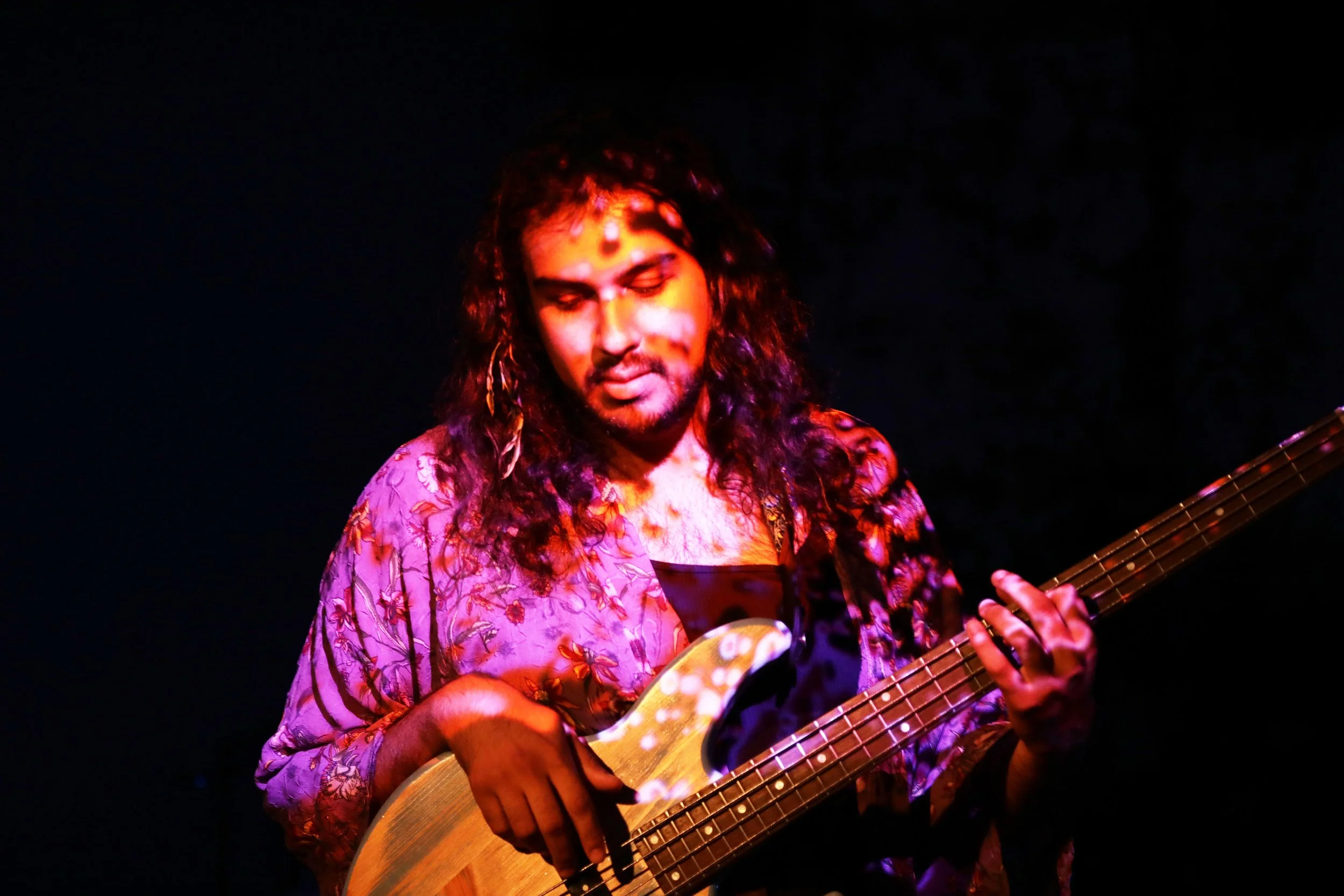 Musician with long curly hair playing an acoustic guitar, wearing a colorful floral shirt, with a dark background.