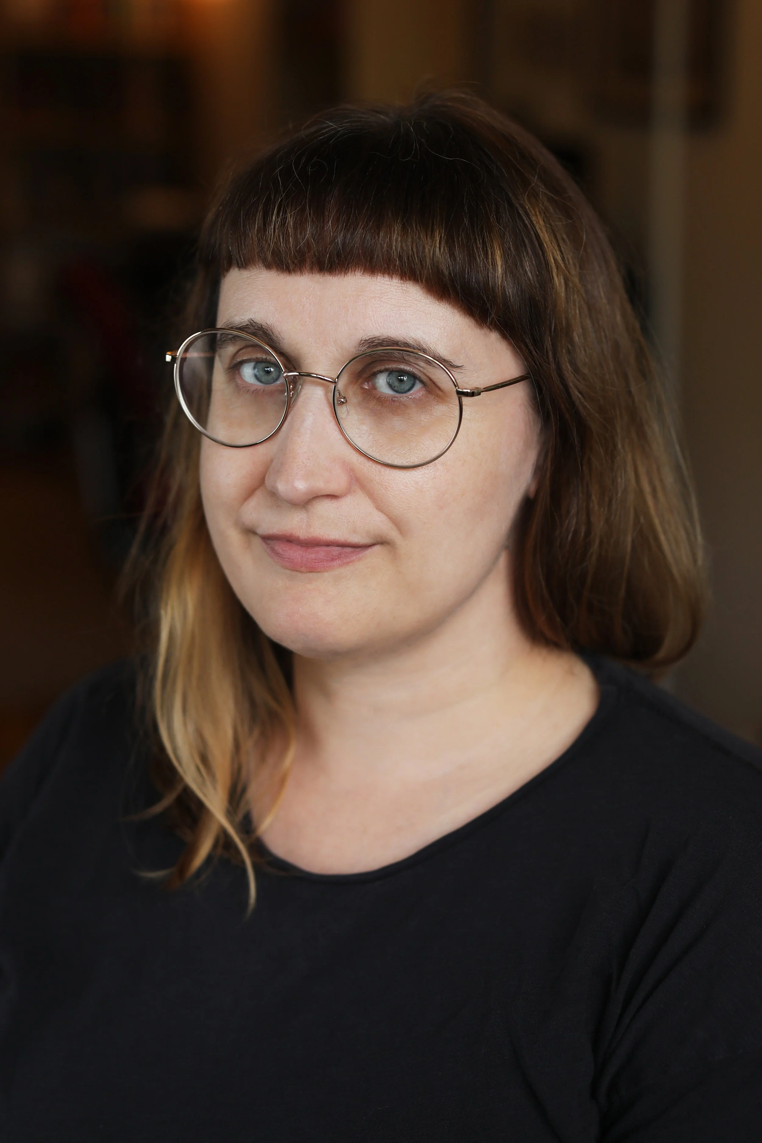 A woman with light skin, brown hair with blonde tips, round glasses, and a black top, looking slightly to the left in a softly lit indoor setting.