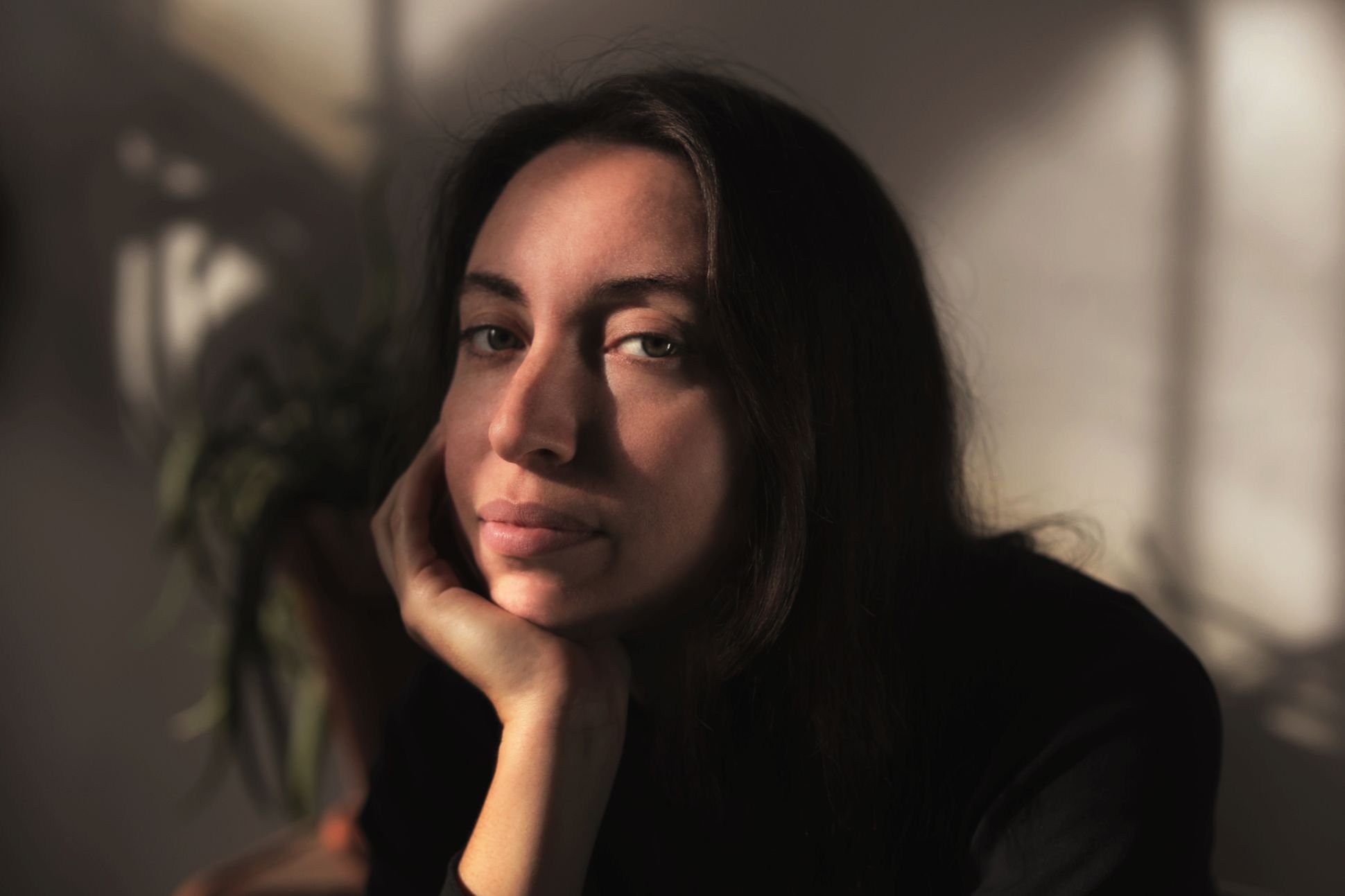 A woman with dark hair and light eyes is resting her face on her hand, looking at the camera with a slight smile.