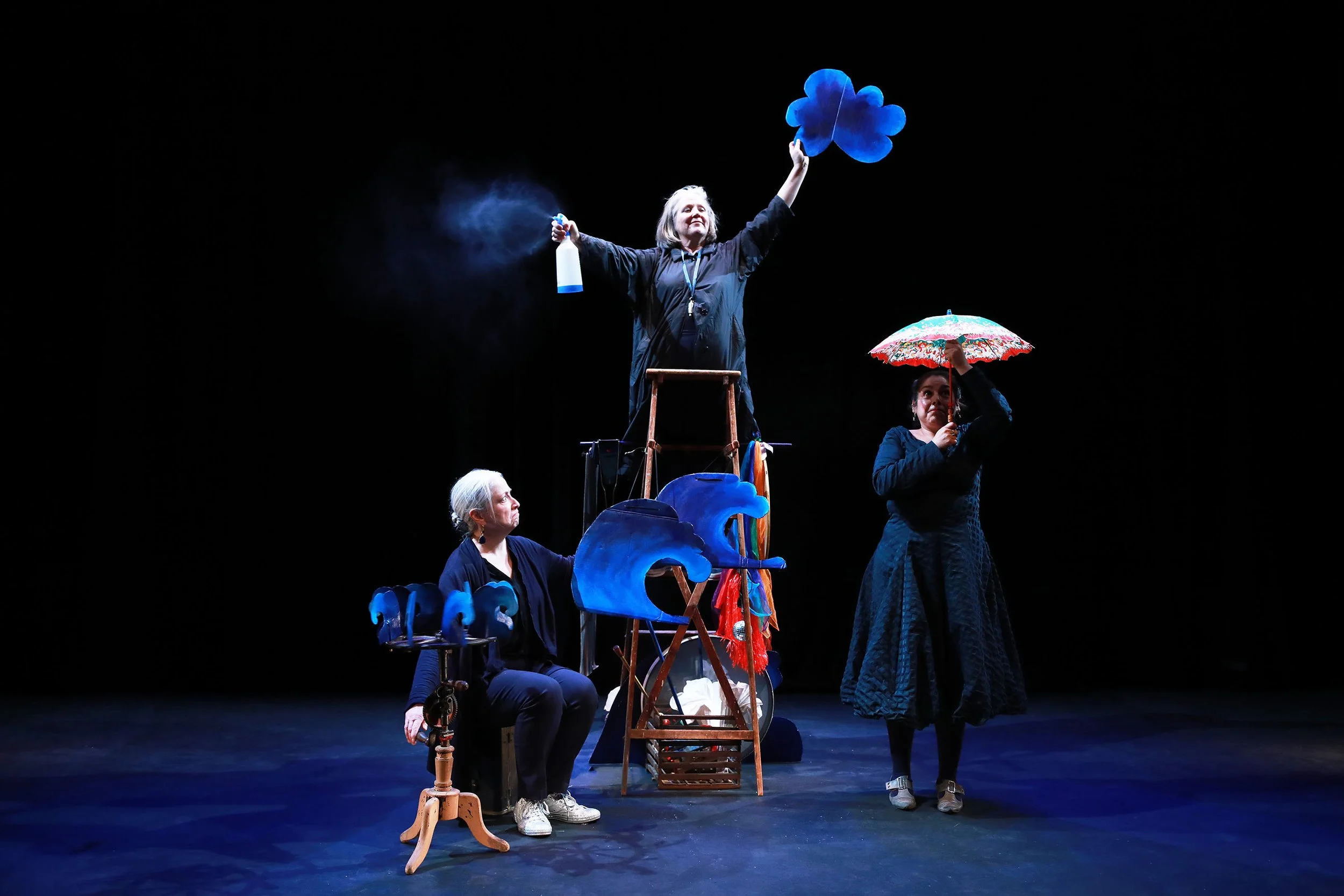 Four women practicing a theatrical performance on stage, with one woman standing on a ladder holding a large blue shamrock-shaped prop, another holding an umbrella, and two others seated or standing in front, all dressed in black against a dark backg