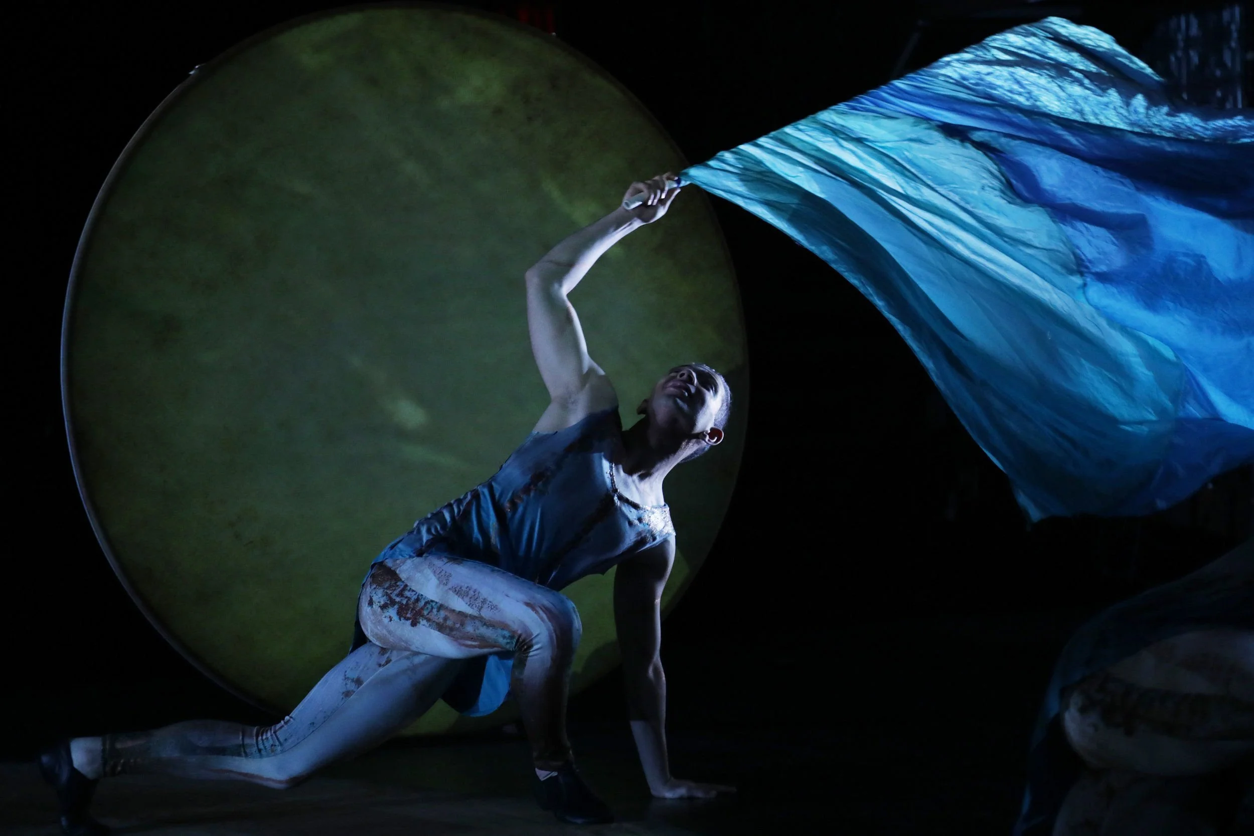 Performer in a black and white costume stretching on stage with a large, illuminated blue fabric flowing overhead.