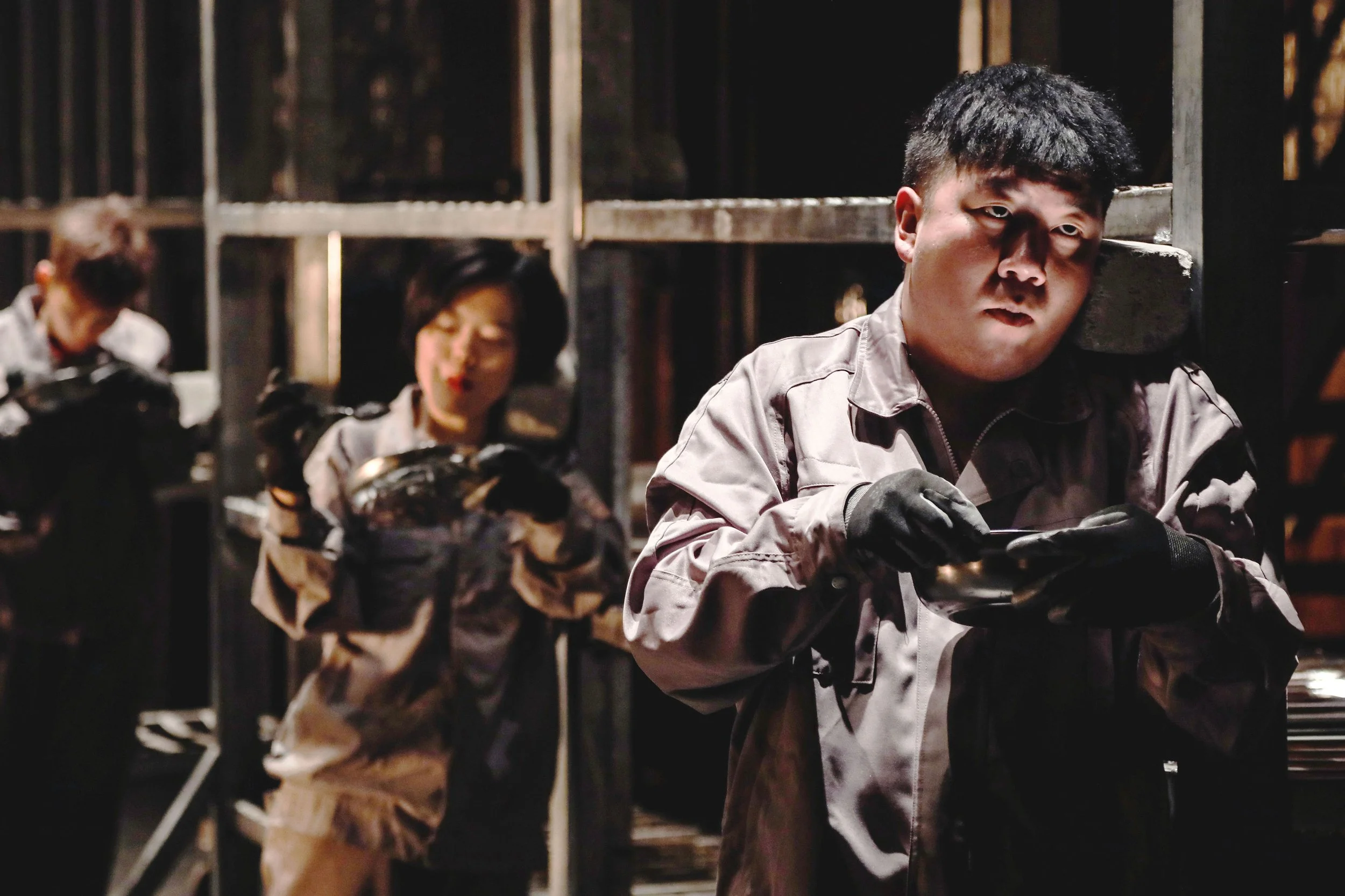 Three individuals in gray uniforms with black gloves working with guns in a dark, industrial environment with metal scaffolding.