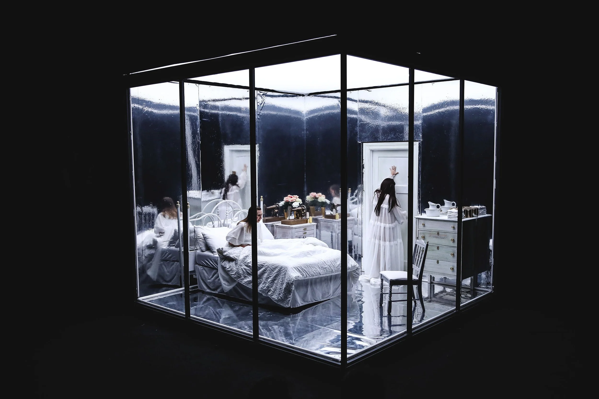 A bedroom enclosed in glass walls with a black ceiling, featuring a white bed, flowers, a white dresser, and a woman in white dress.